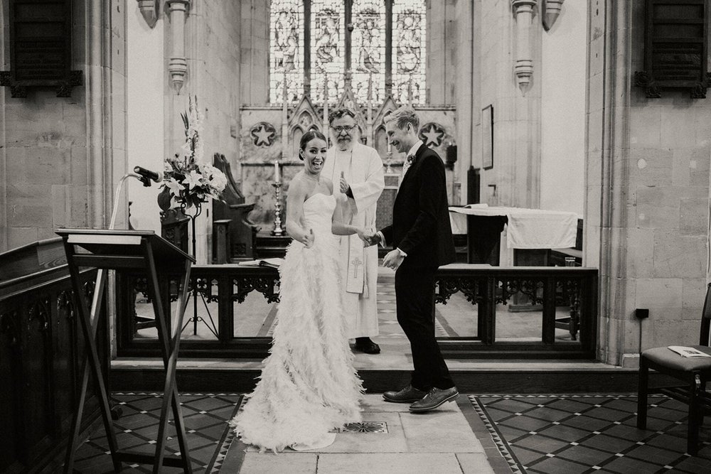  A black and white image of Sophie and Pete at the front of the church for the ceremony. Sophie has turned to the camera smiling and Pete looks at Sophie.  