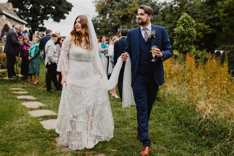  Gemma and Nick down a path surrounded by a lawned area. Nick holds Gemma’s veil off the ground as they walk. 