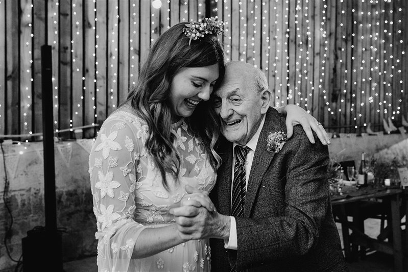  Gemma and an elderly man stand hand in hand whilst embracing one another, in front of the barn’s timber walls covered in fairy lights. 