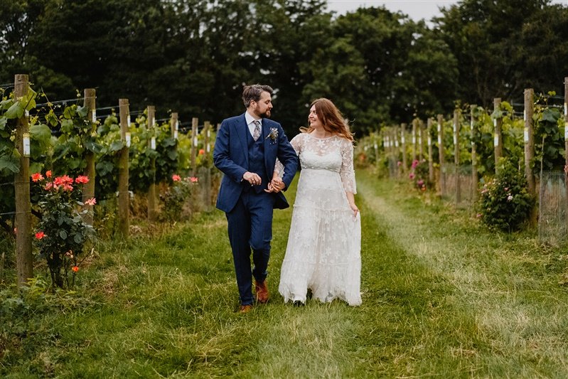  Nick and Gemma walk between rows of vegetation holding hands and looking at one another. Nick wears a navy blue suit. 