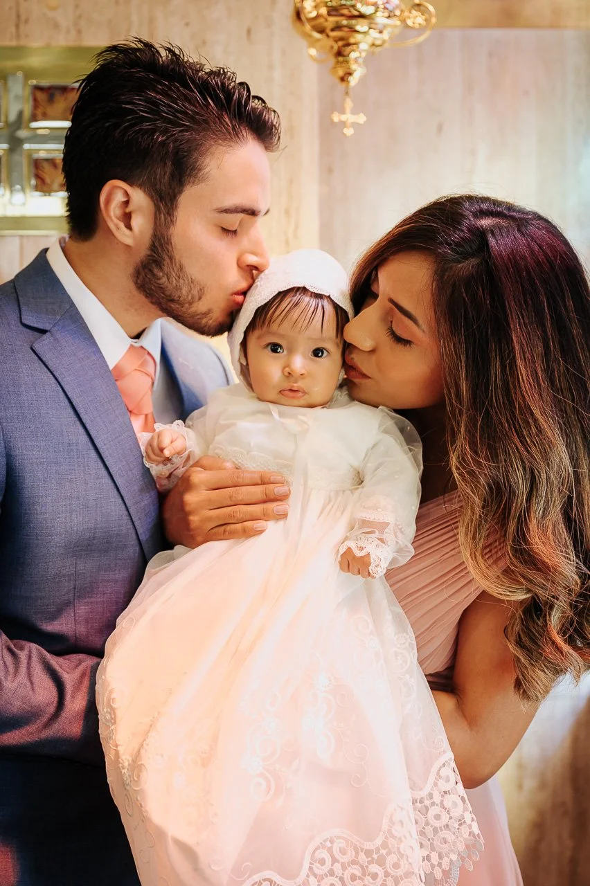 Una pareja sostiene a su bebé en un bautizo, la pareja la besa en la frente y ella mira hacia la cámara, en un ambiente elegante con una lámpara de oro en el fondo.