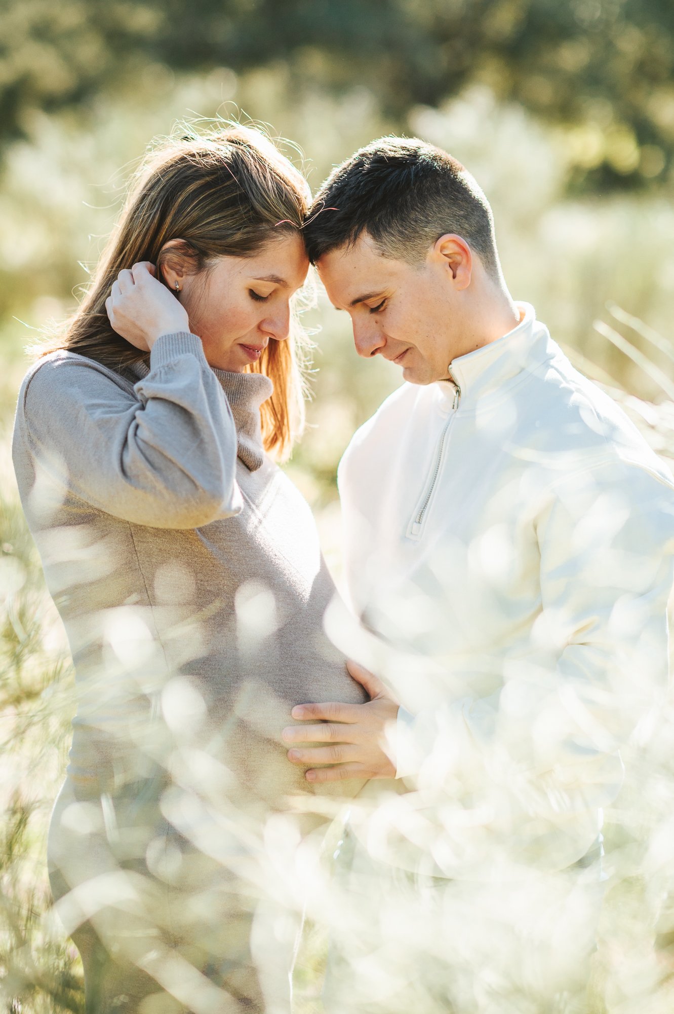 Una pareja frente a frente, con manos sobre un vientre de embarazo, en un entorno natural, ambos con expresiones de felicidad y ternura.