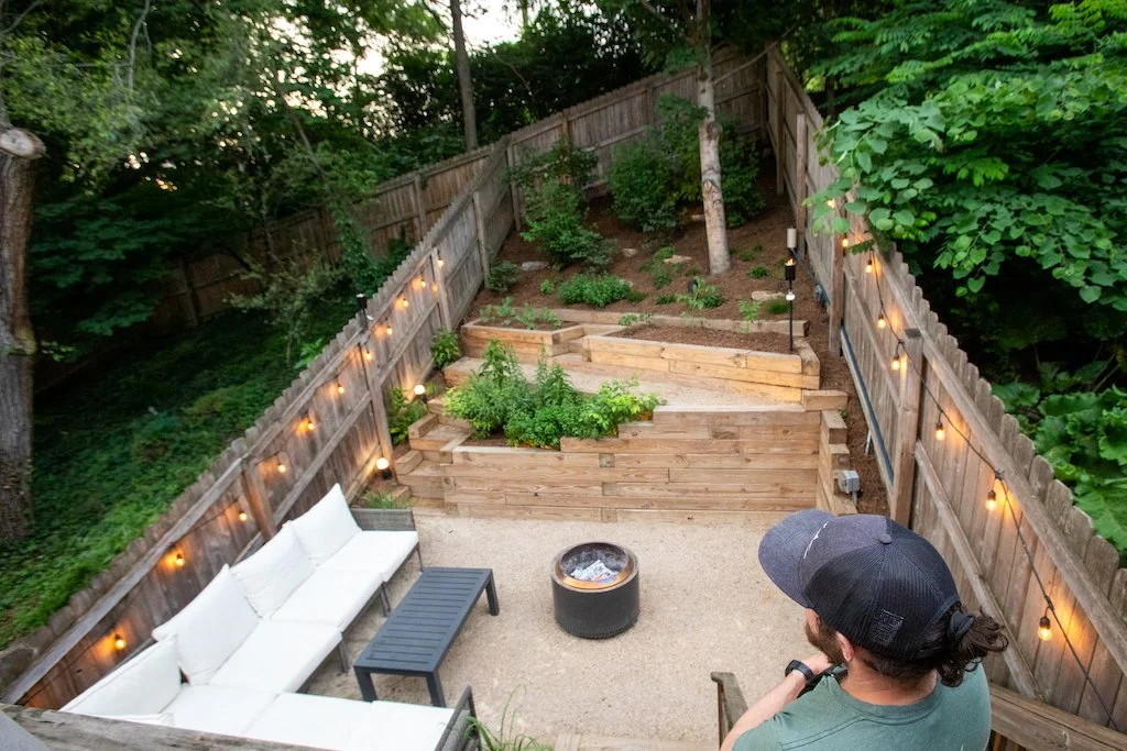 John Hemmerle, founder of Our Land Organics, overlooks a background landscaped with a crushed stone fire pit, wooden terraced garden beds, and native plants