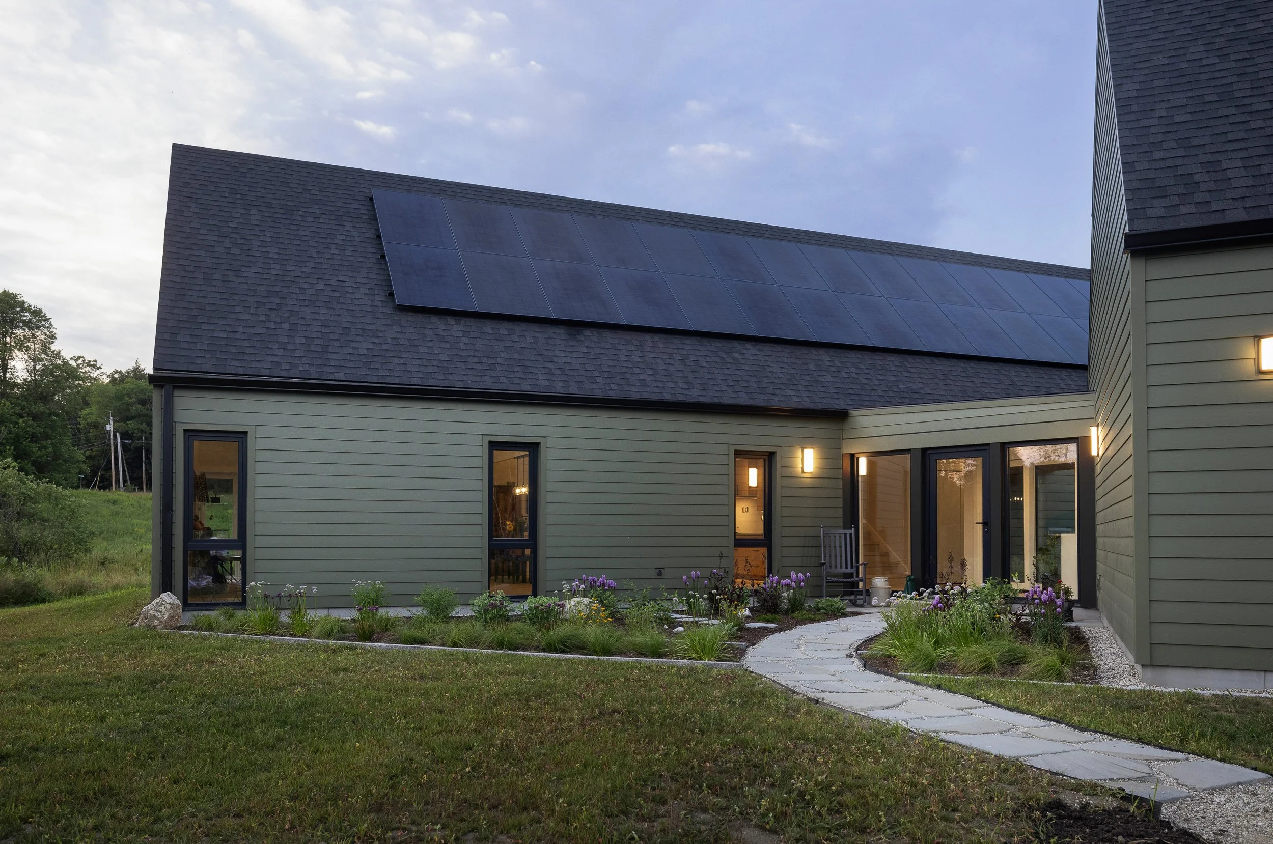 A modern house with solar panels on the roof, green siding, and a curved stone pathway leading to the entrance, surrounded by a garden with plants and flowers, during the evening.