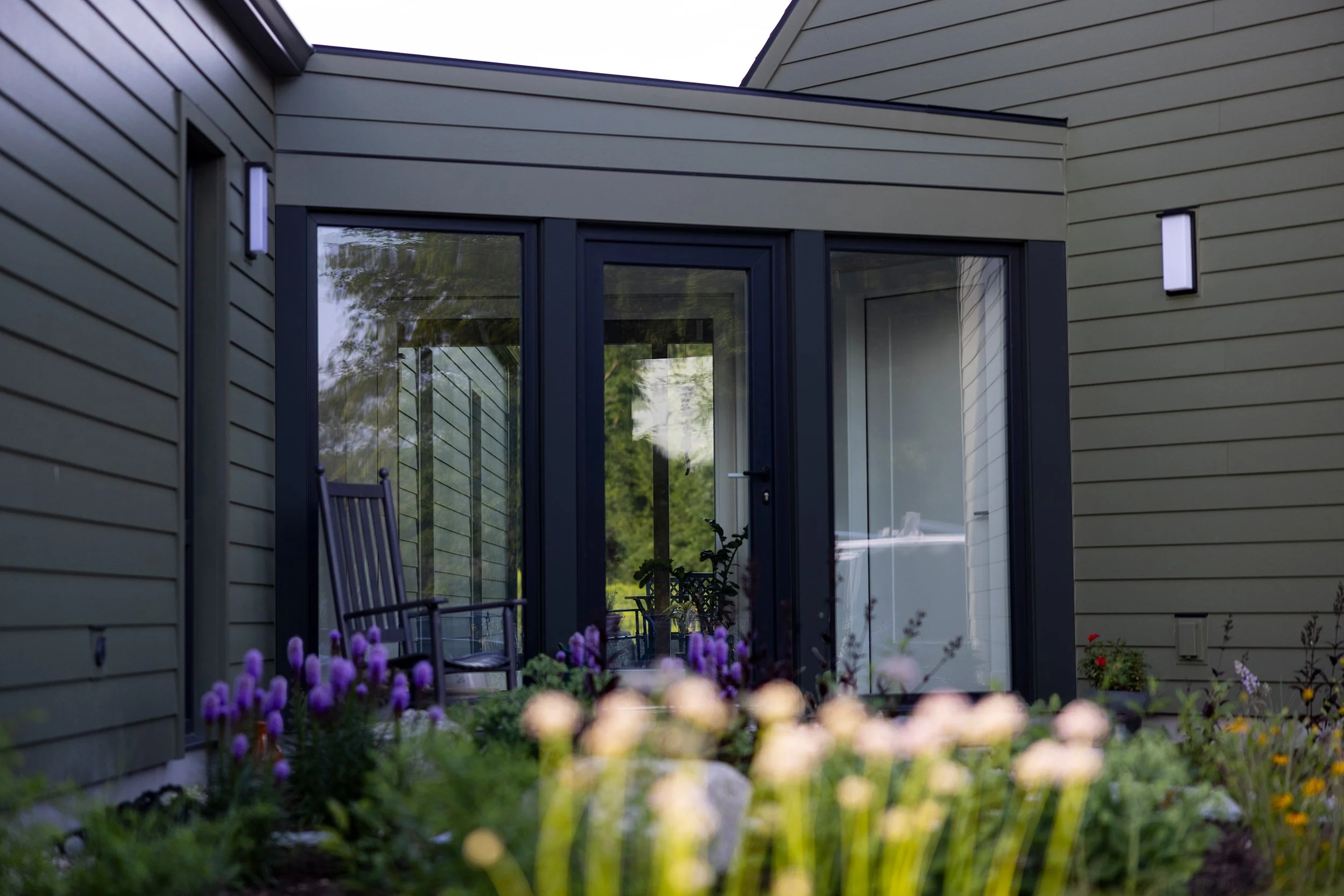 A modern house with green siding and large glass sliding doors, a porch with chairs, and a garden with purple and yellow flowers.
