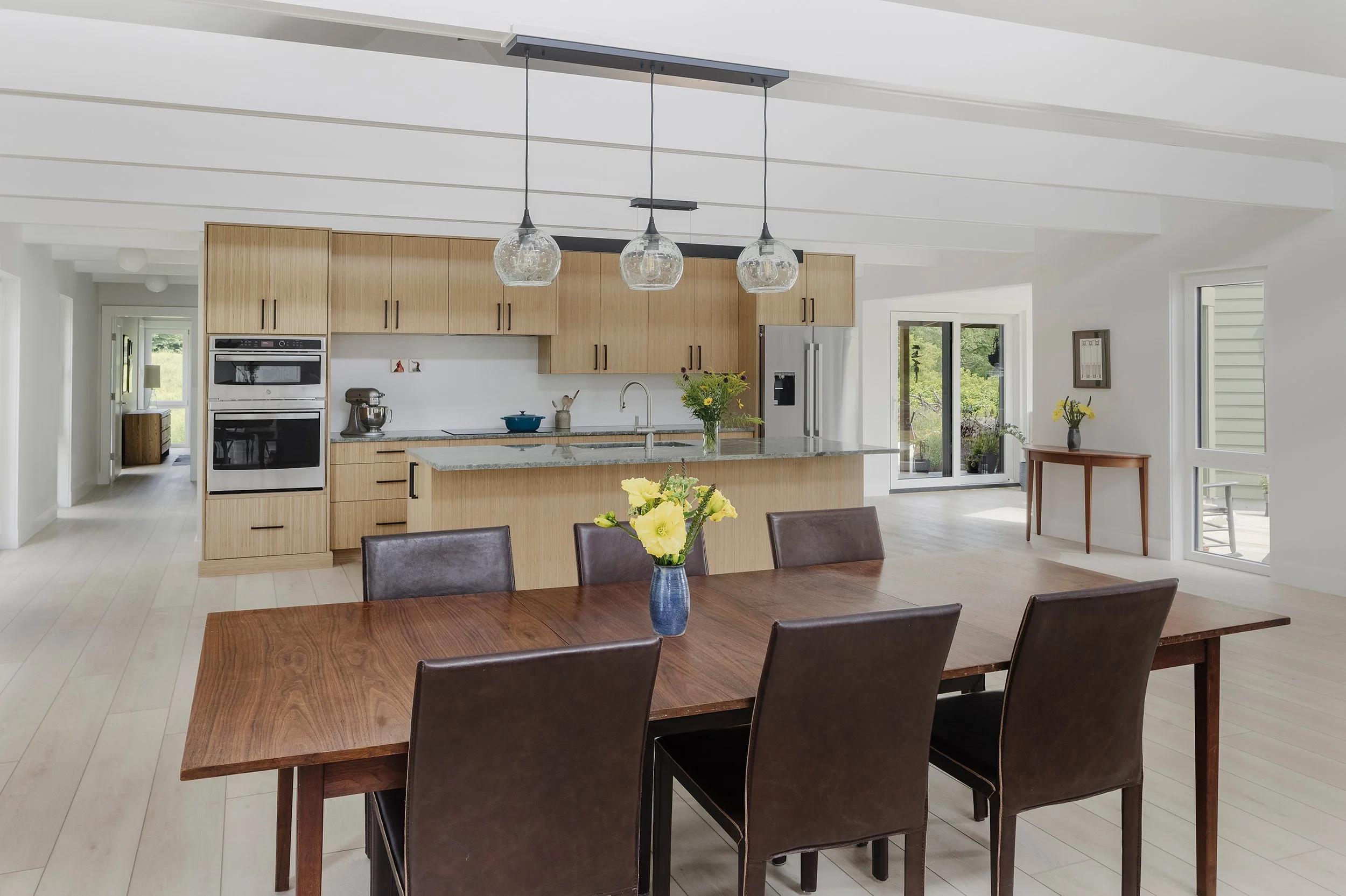 Modern open-plan kitchen with a large wooden dining table and six brown leather chairs, a kitchen island with a granite countertop, hanging pendant lights, wooden cabinets, stainless steel appliances, and large windows with a view of greenery outside