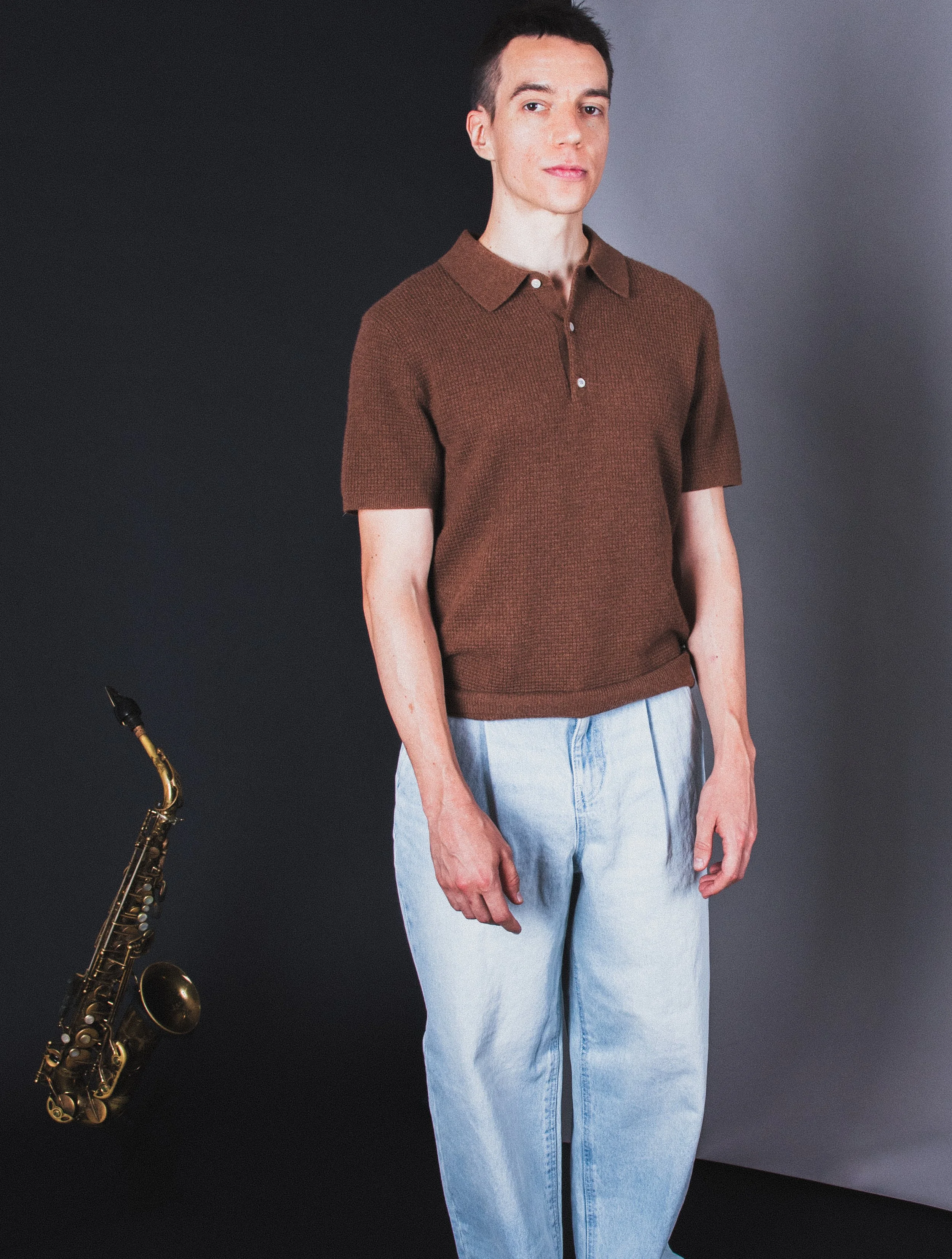 A person with short dark hair wearing a brown short-sleeved polo shirt and light blue jeans standing against a dark background. Part of a saxophone is visible to the left.