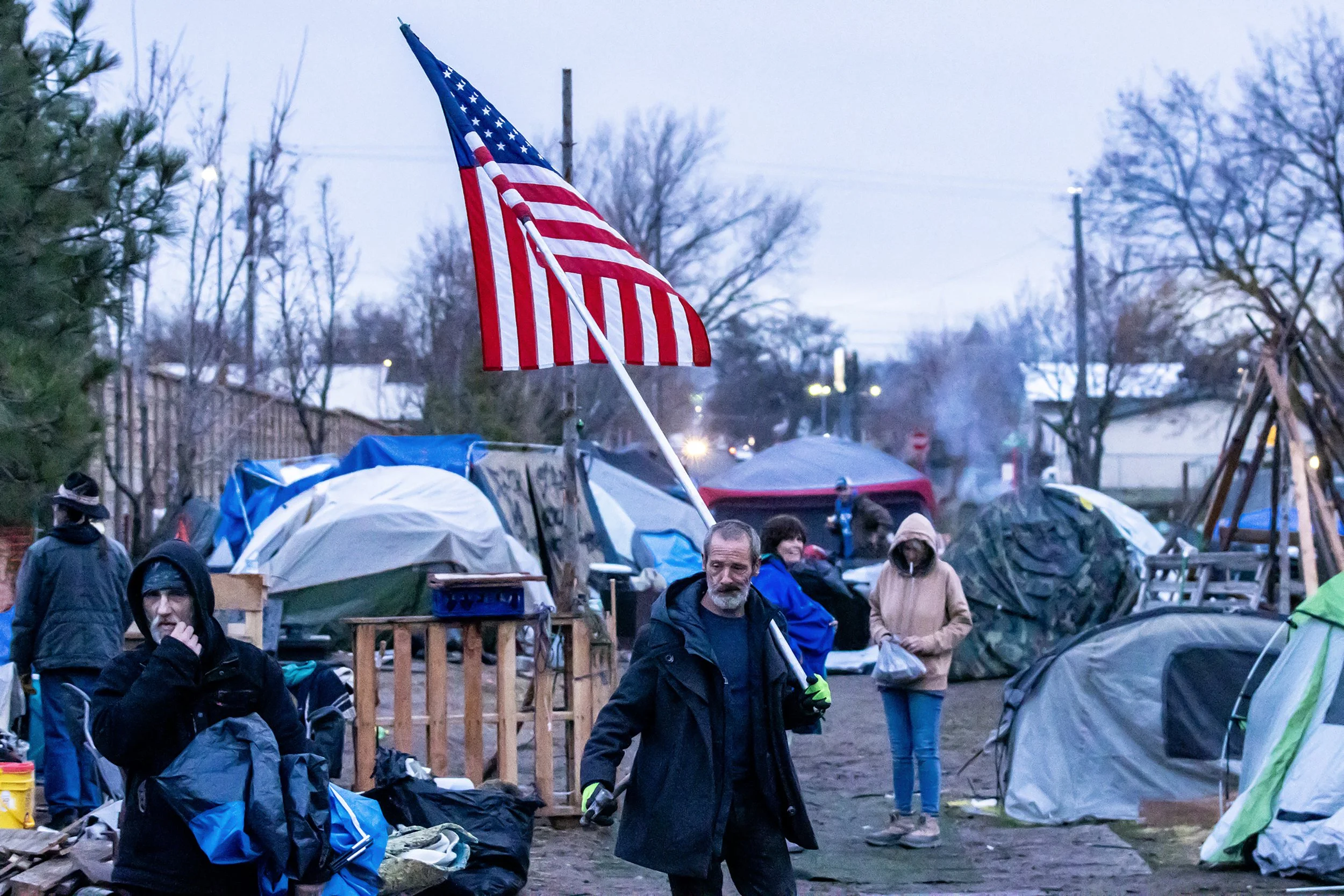  A camp resident walks through the camp with an American flag as residents work to gather up their belongings and move to a new area Monday, Feb. 12, 2024, in Clarkston. Efforts by volunteers and residents to clean up the camp moved late into the nig