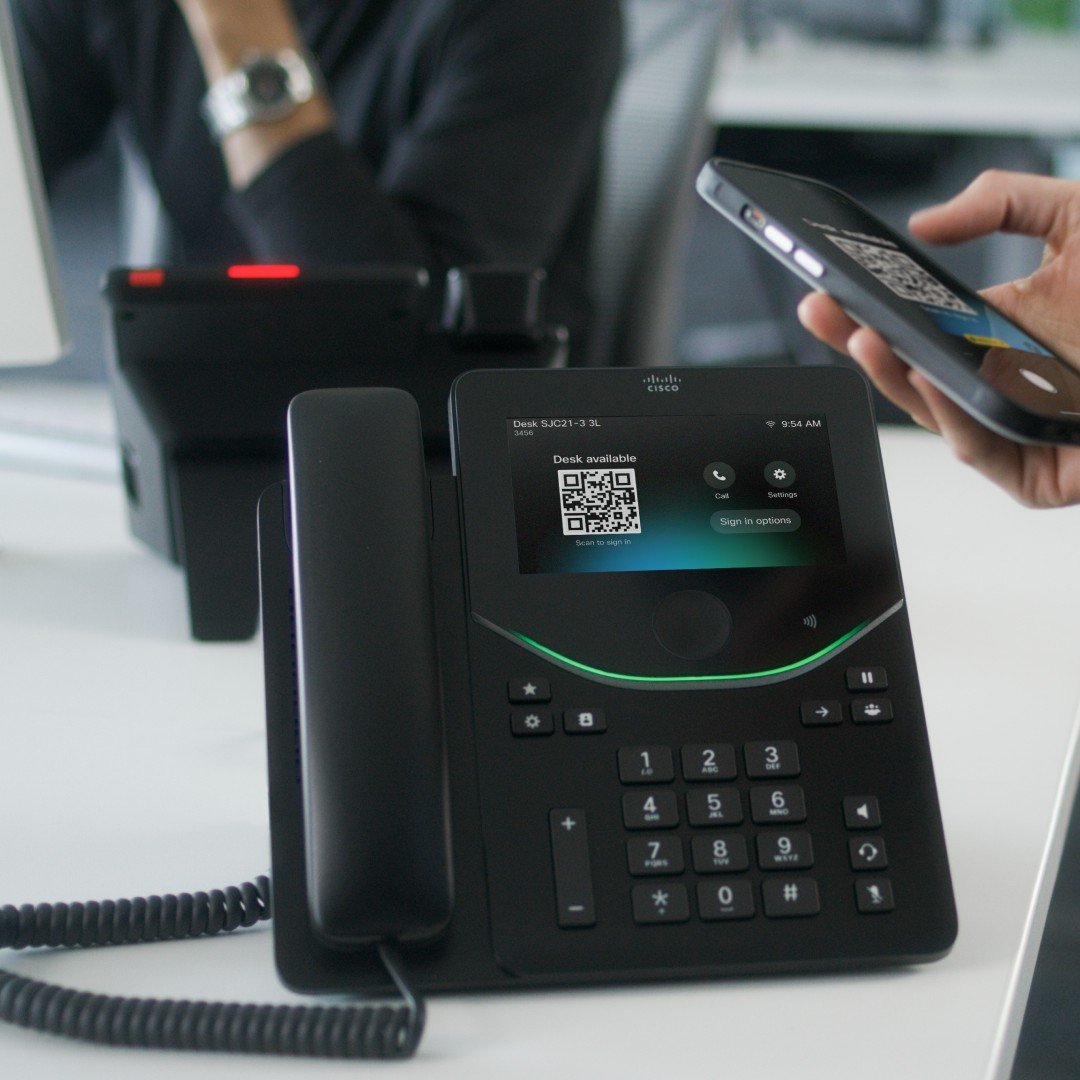 Person checking in to a shared desk by holding a smartphone near a Cisco Desk Phone 9871 that shows a desk-available QR code