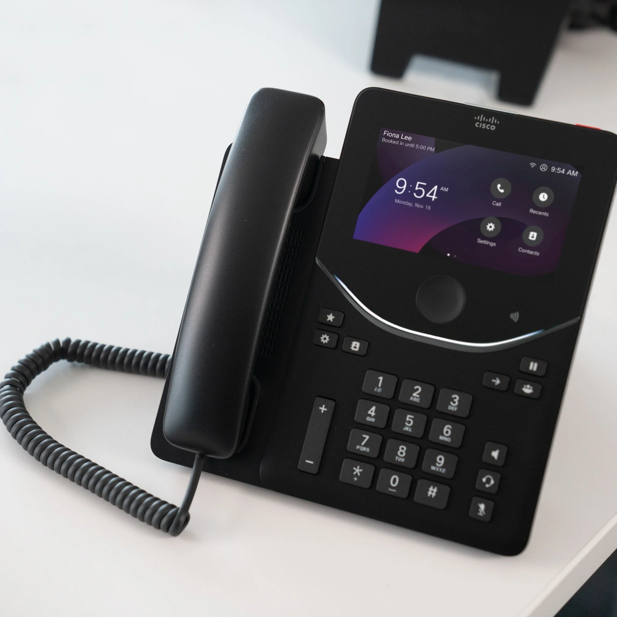 Cisco Desk Phone 9871 on an office desk, showing a booked-desk greeting and call, recents, settings and contacts icons on its touchscreen