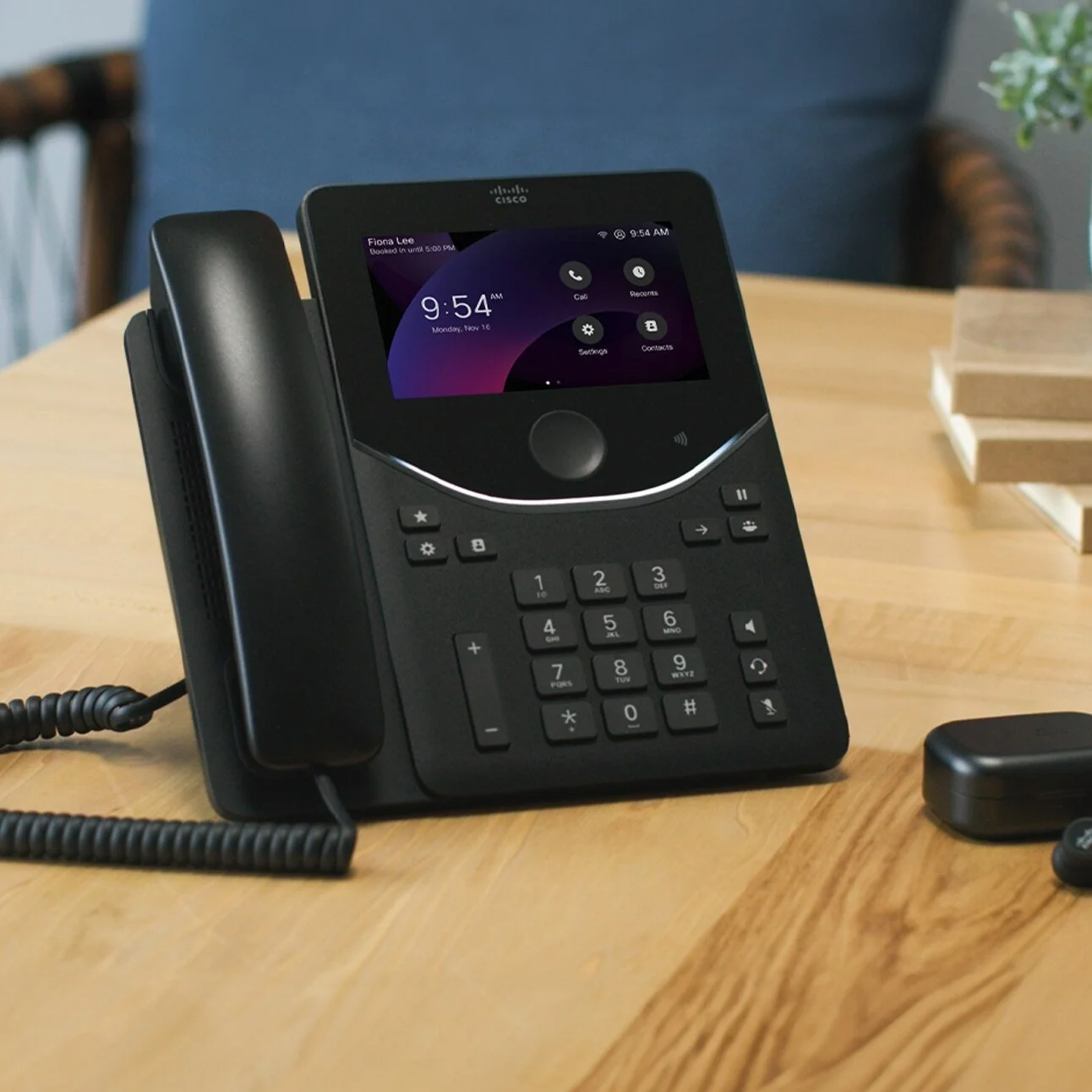 Cisco Desk Phone 9871 in Carbon Black on a wooden desk with its touchscreen home screen lit