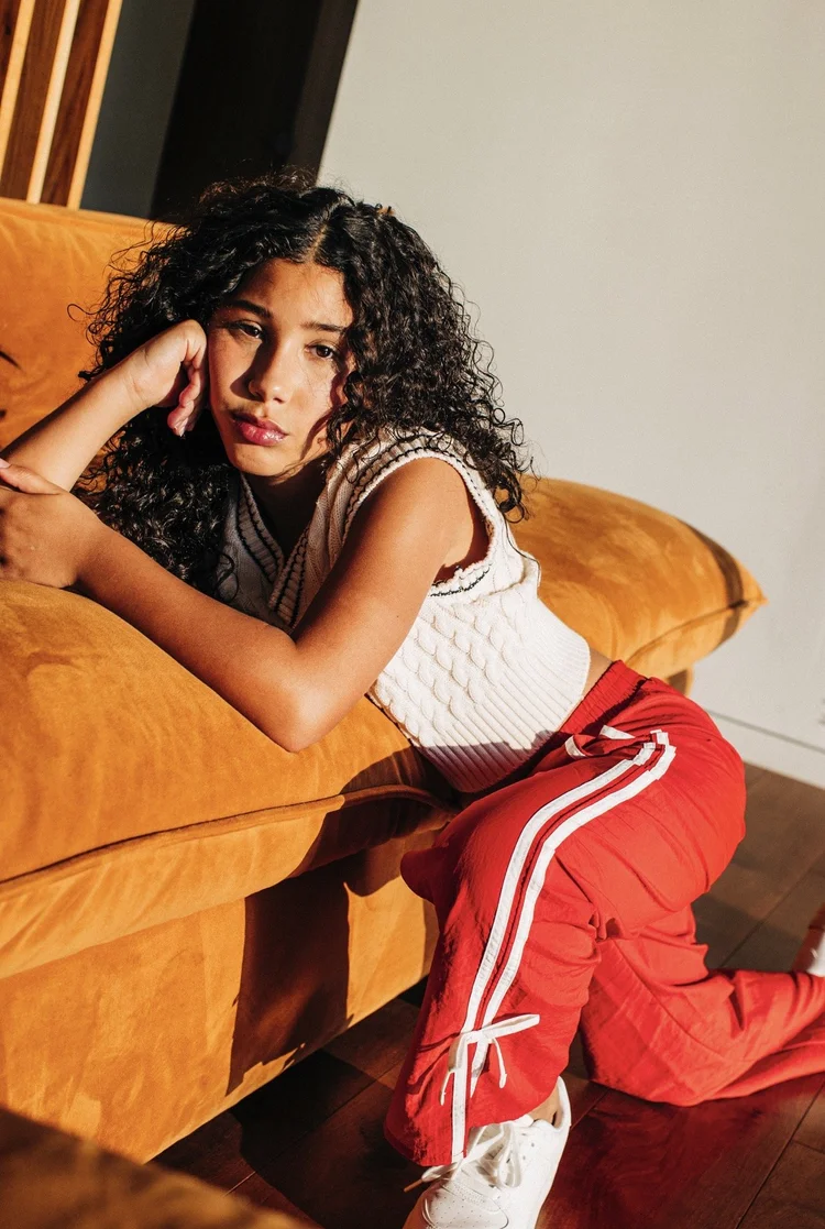 Young model with natural curls seated on an orange sofa in red athletic trousers