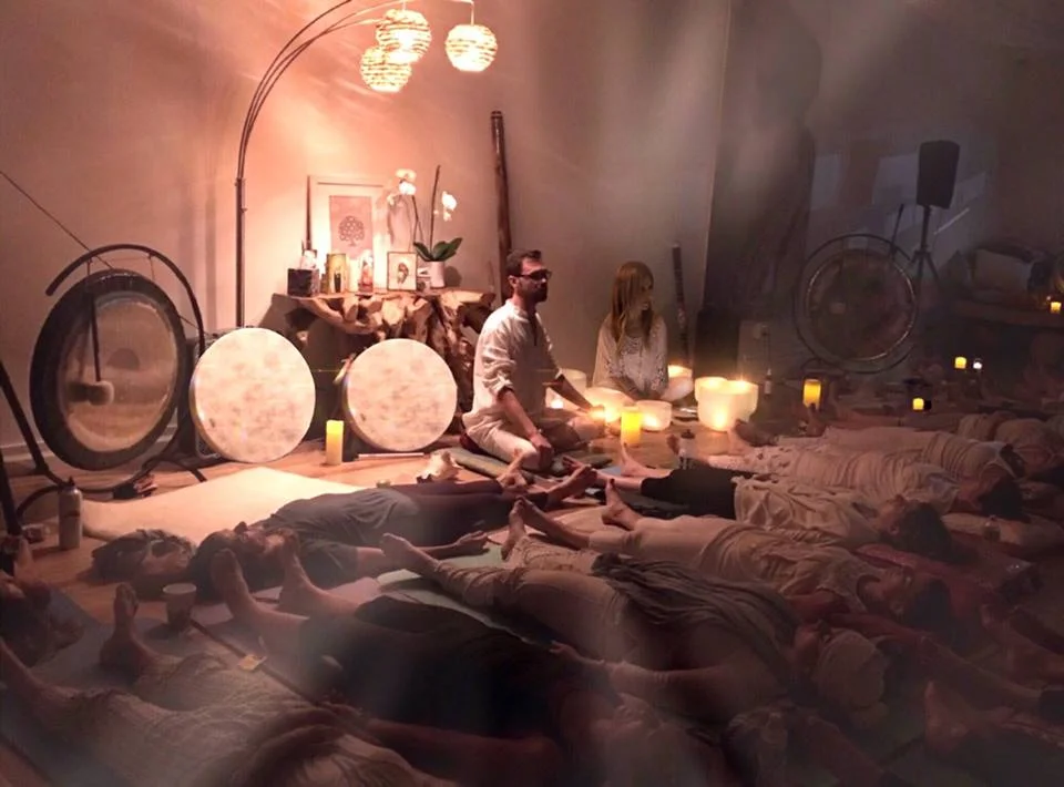 A serene meditation room with dim lighting, featuring people lying on mats. Instruments and candles set a calming atmosphere. Warm tones convey peace.