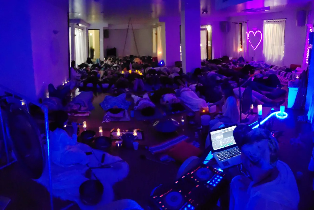 A dimly lit room with purple and blue lighting, filled with people lying on mats. Soft candlelight and a neon heart lend a calm, serene ambiance.