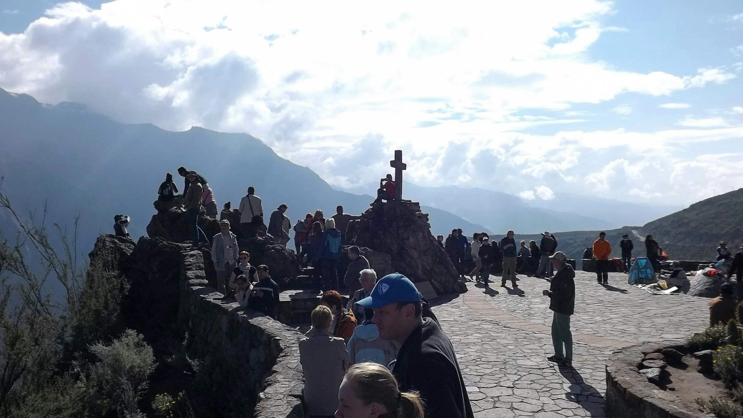 A large group of people gathered on a stone terrace at the edge of a mountainous landscape, with a cross monument and a bright, cloudy sky above.