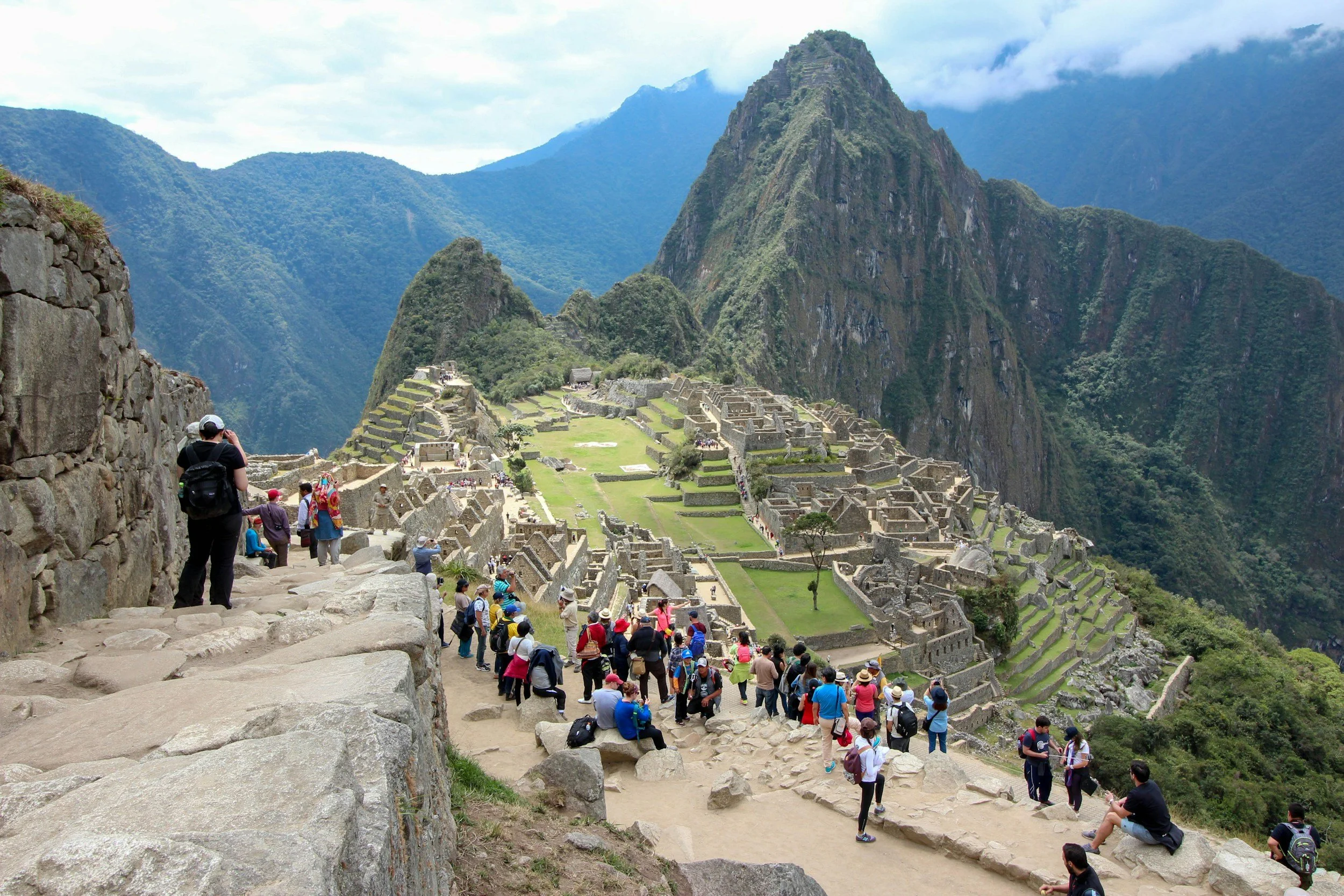A group of tourists explore the ancient ruins of Machu Picchu, set against lush green mountains under a partially cloudy sky. The scene conveys awe.