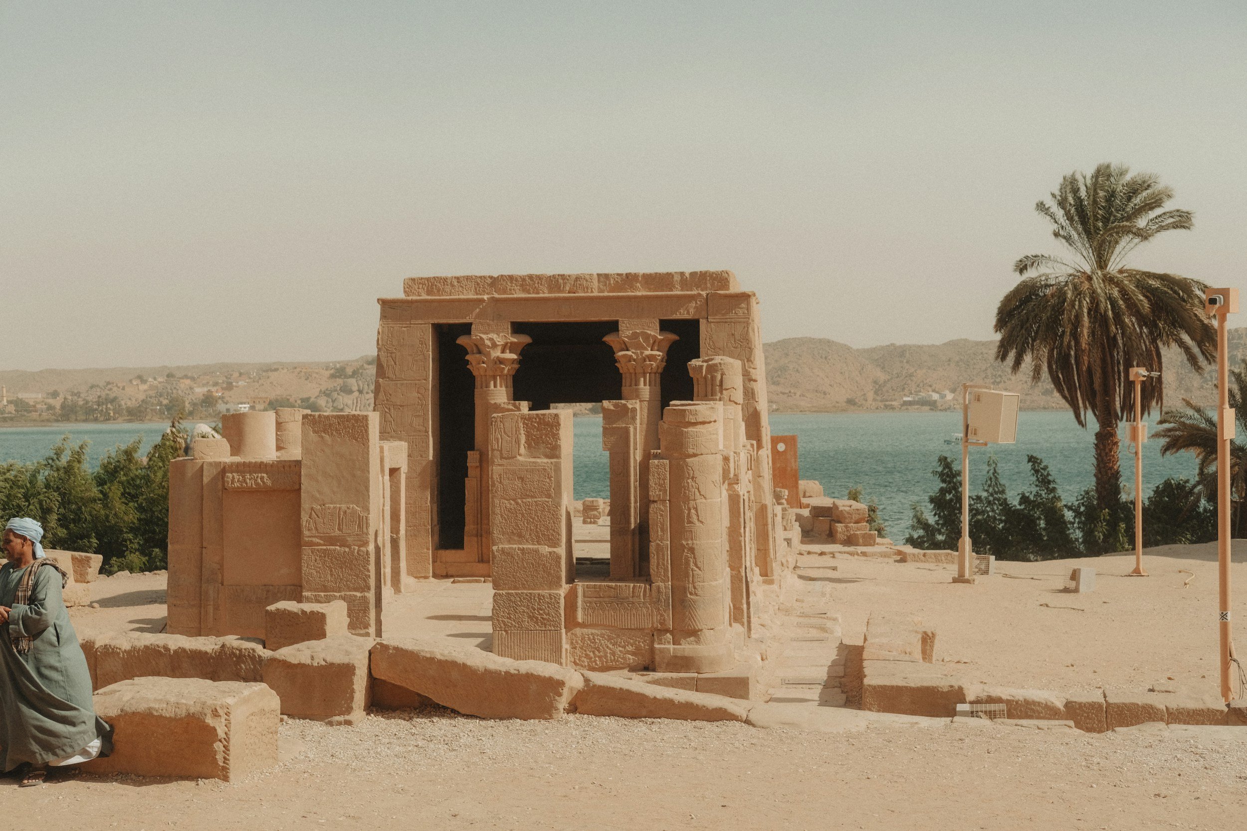 Ancient stone ruins sit on sandy terrain with a backdrop of a river and distant hills. A lone palm tree stands nearby, evoking a serene atmosphere.