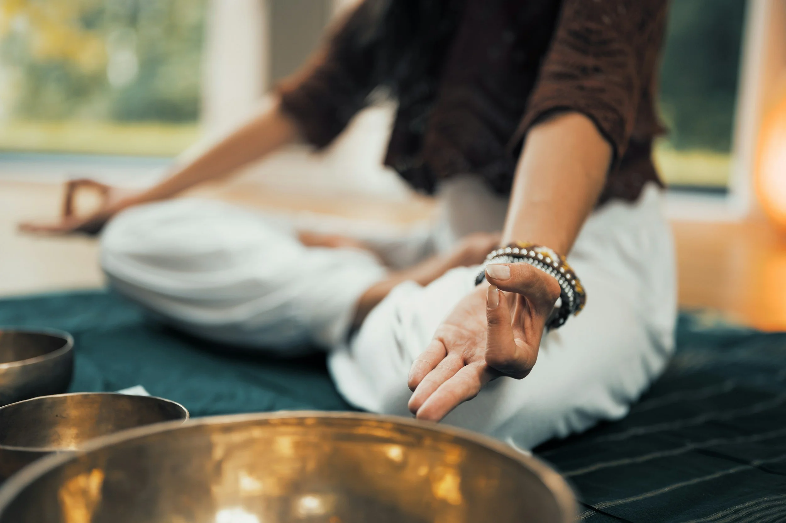 A person sits cross-legged on a mat, wearing a beaded bracelet, with an open palm facing up. Meditation bowls are nearby, evoking calm and focus.