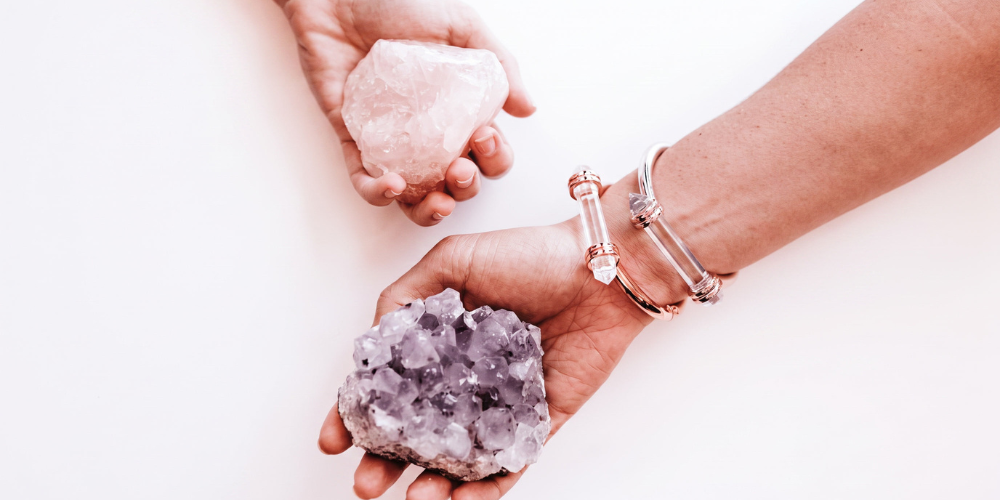 Two hands hold raw crystals: a pale pink stone above and a purple amethyst cluster below, with bracelets visible against a white background.
