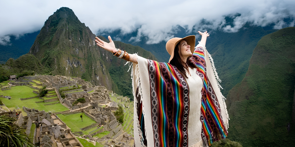 Joyful woman in a colorful Andean poncho and hat raises her arms at Machu Picchu, with ancient ruins, green mountains, and clouds behind her.