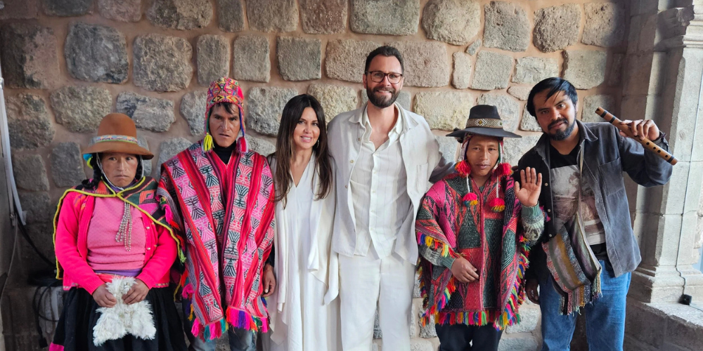 Group portrait of five people in colorful Andean clothing and two guests outside a stone wall, smiling warmly in a festive cultural setting.