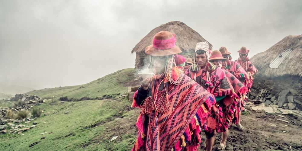 Indigenous Andean villagers in vivid pink hats and woven ponchos walk through a misty mountain village, led by a smoking elder.