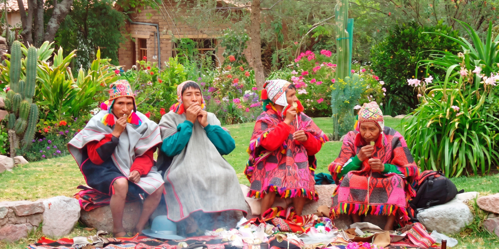 Four Indigenous people in colorful traditional clothing sit around a smoking fire in a lush garden, creating a vibrant, contemplative scene.