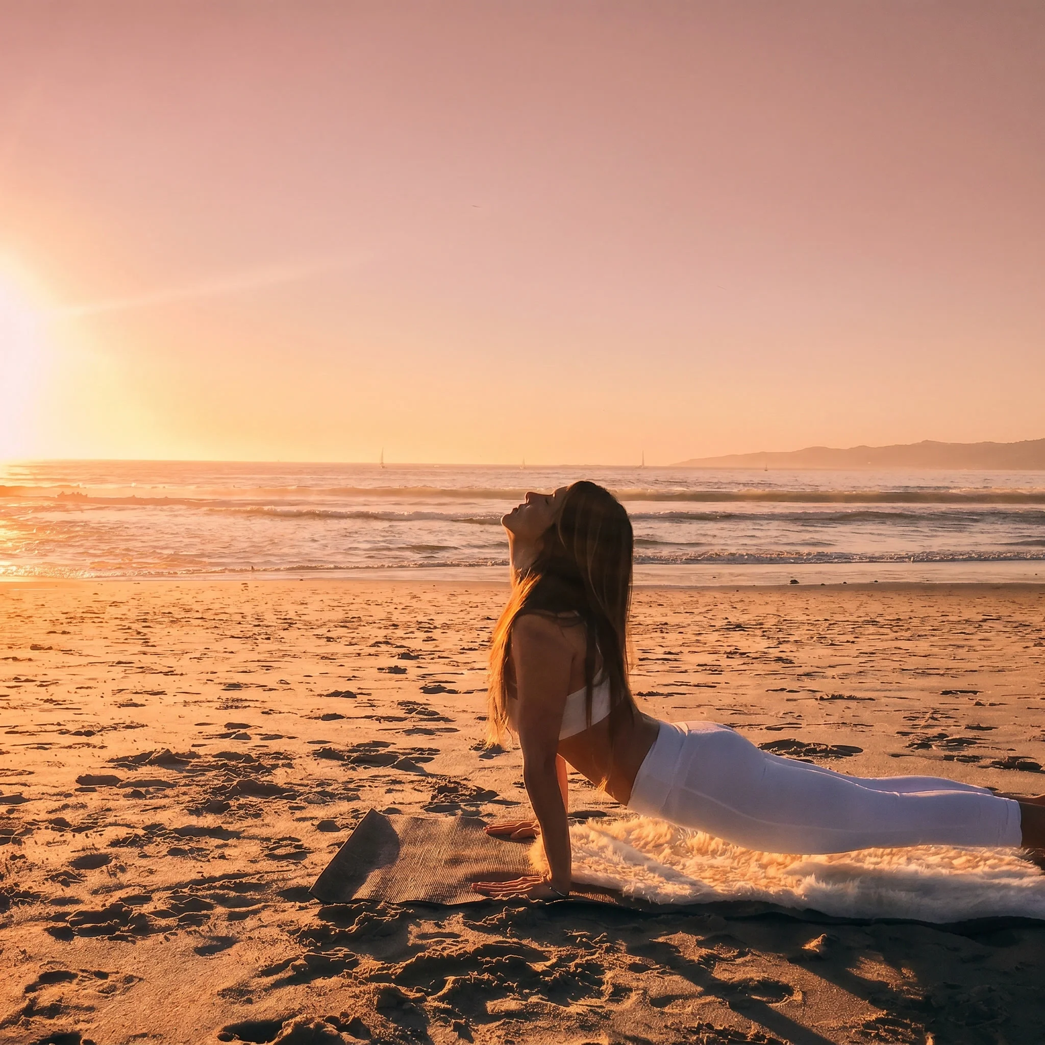 A person practices yoga in a cobra pose on a sandy beach at sunset. The warm glow of the sun and calm ocean create a serene, peaceful atmosphere.