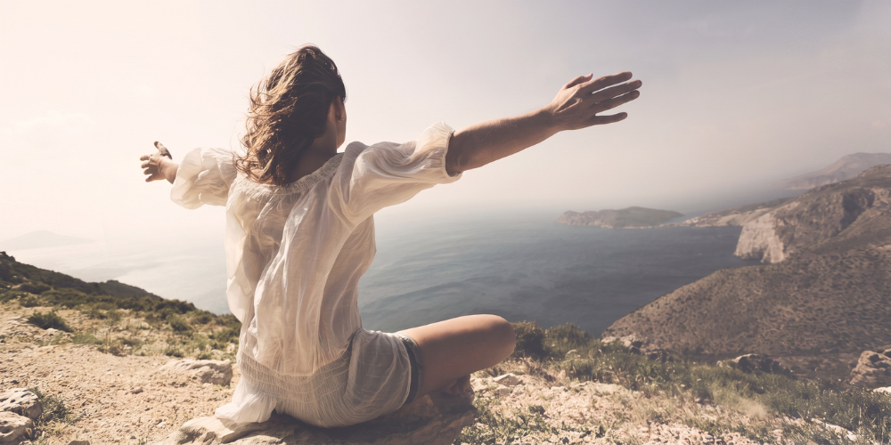 Woman practicing mindful meditation outdoors with arms extended overlooking a scenic coastal landscape.