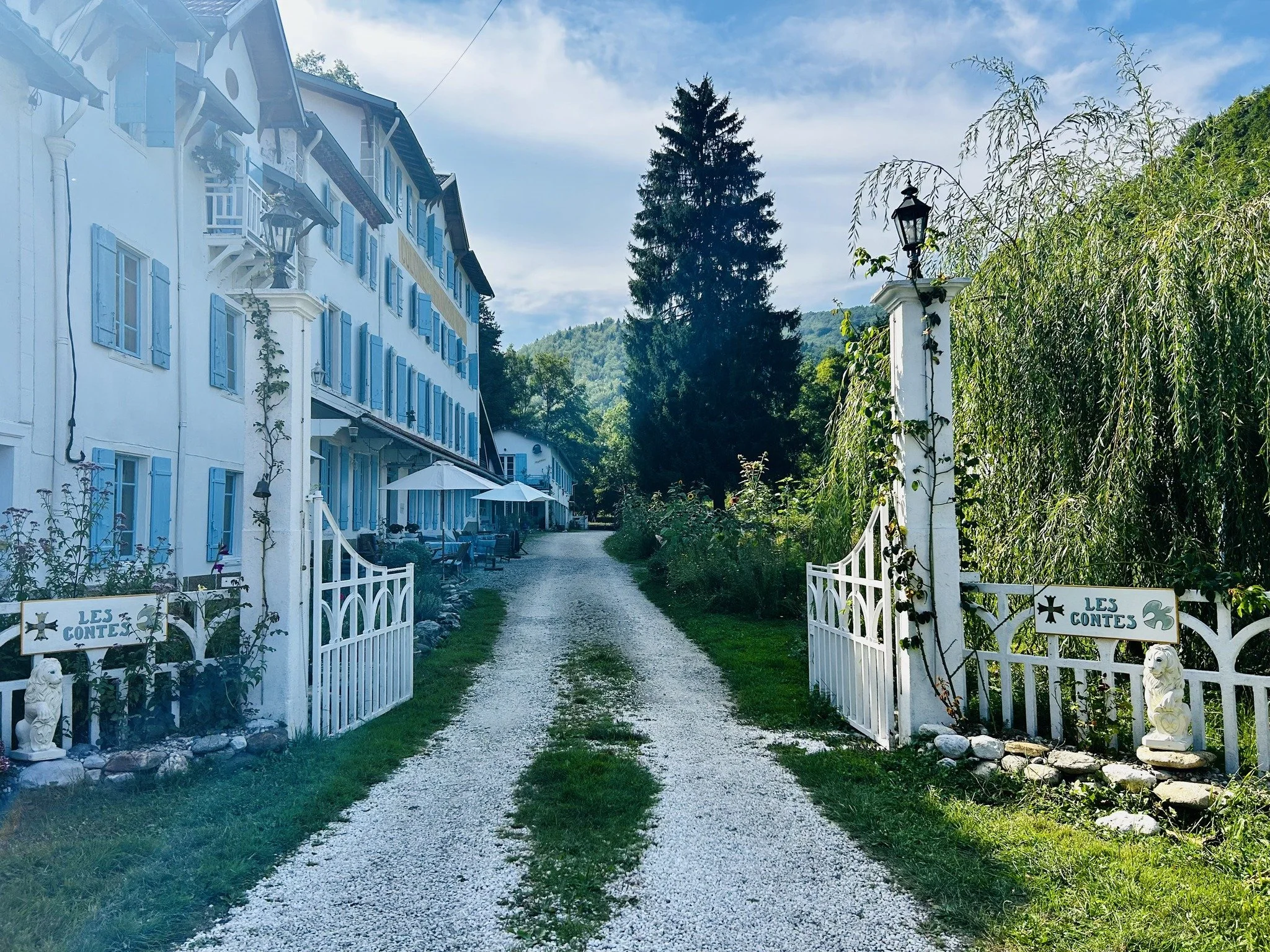 A white gravel path leads to a quaint building with blue shutters on the left, flanked by white gates adorned with climbing plants, under a clear blue sky