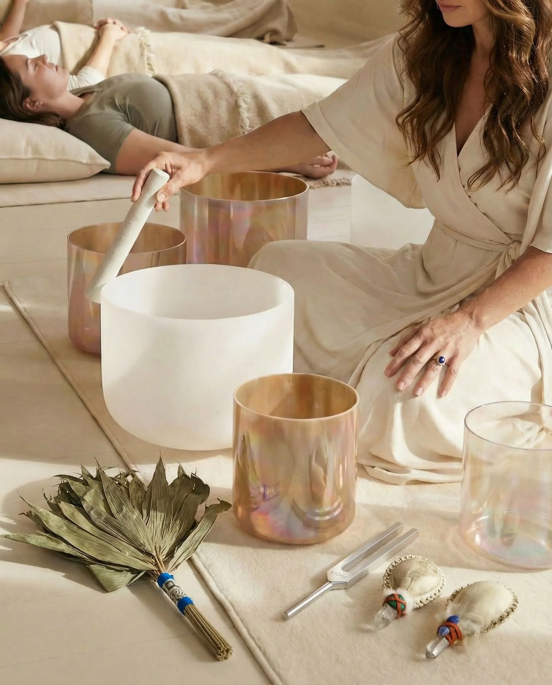 A woman uses a mallet to play crystal singing bowls in a serene setting. A person lies relaxed on a mat in the background, with sage and feathers nearby.