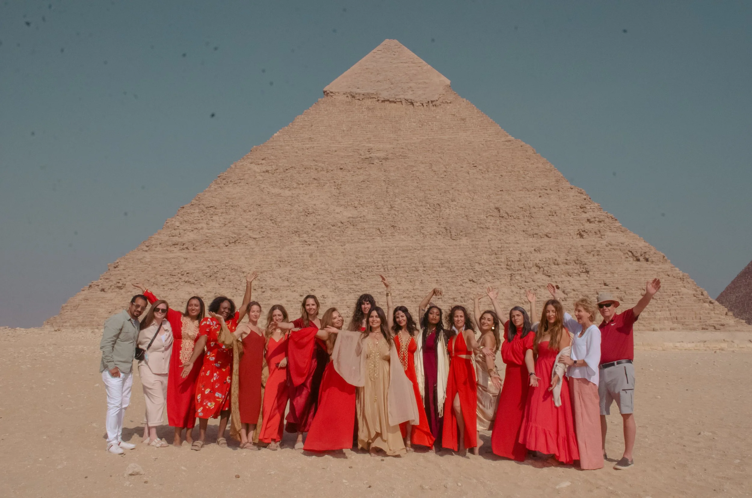 A group of people dressed in red and beige stand joyfully in front of a pyramid under a clear sky, conveying a sense of unity and celebration.