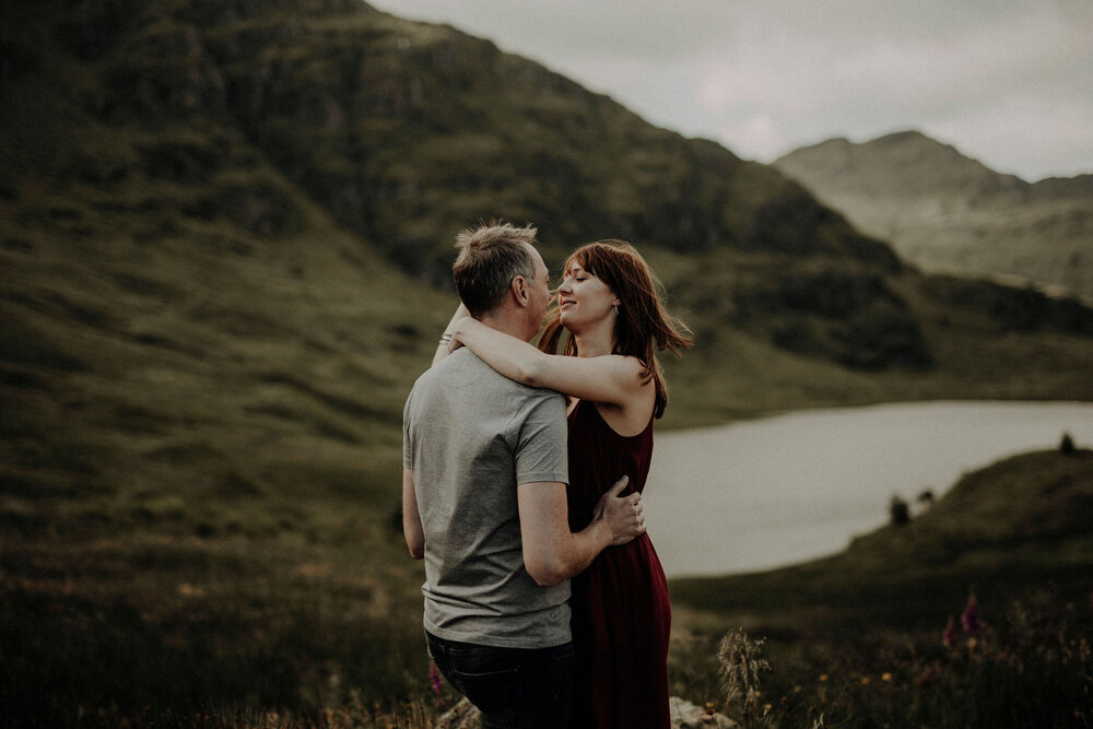  woman in dark red dress and man holding each other in the mountains lake in the back 