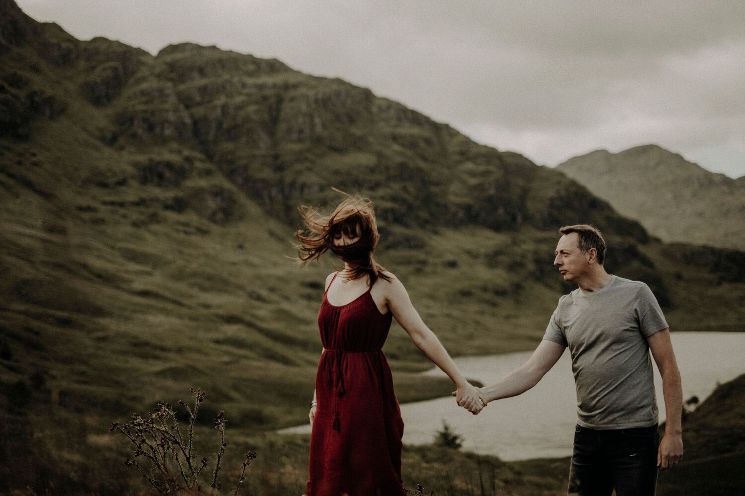  woman and man holding their hands wind blowing hair in mountains with a lake behind 