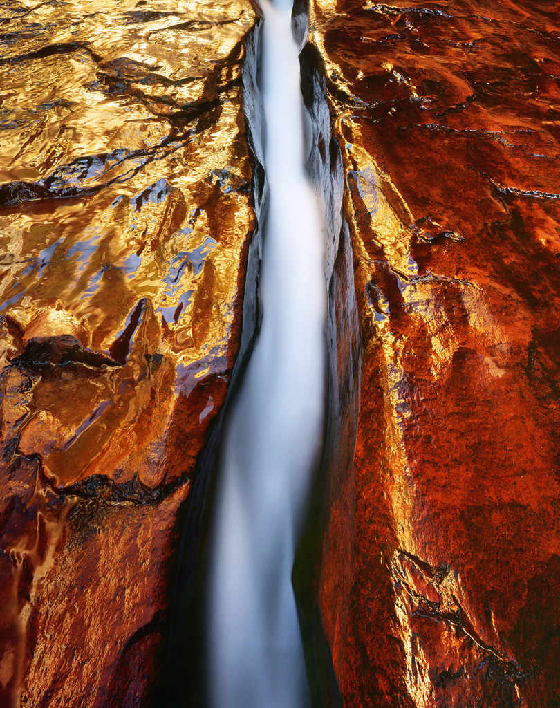 "Sandstone Flow" - An entire creek rushes through a narrow crack in the deep red sandstone. Sunlight reflected on a canyon wall makes the whole image glow brilliantly. Prints Available.
