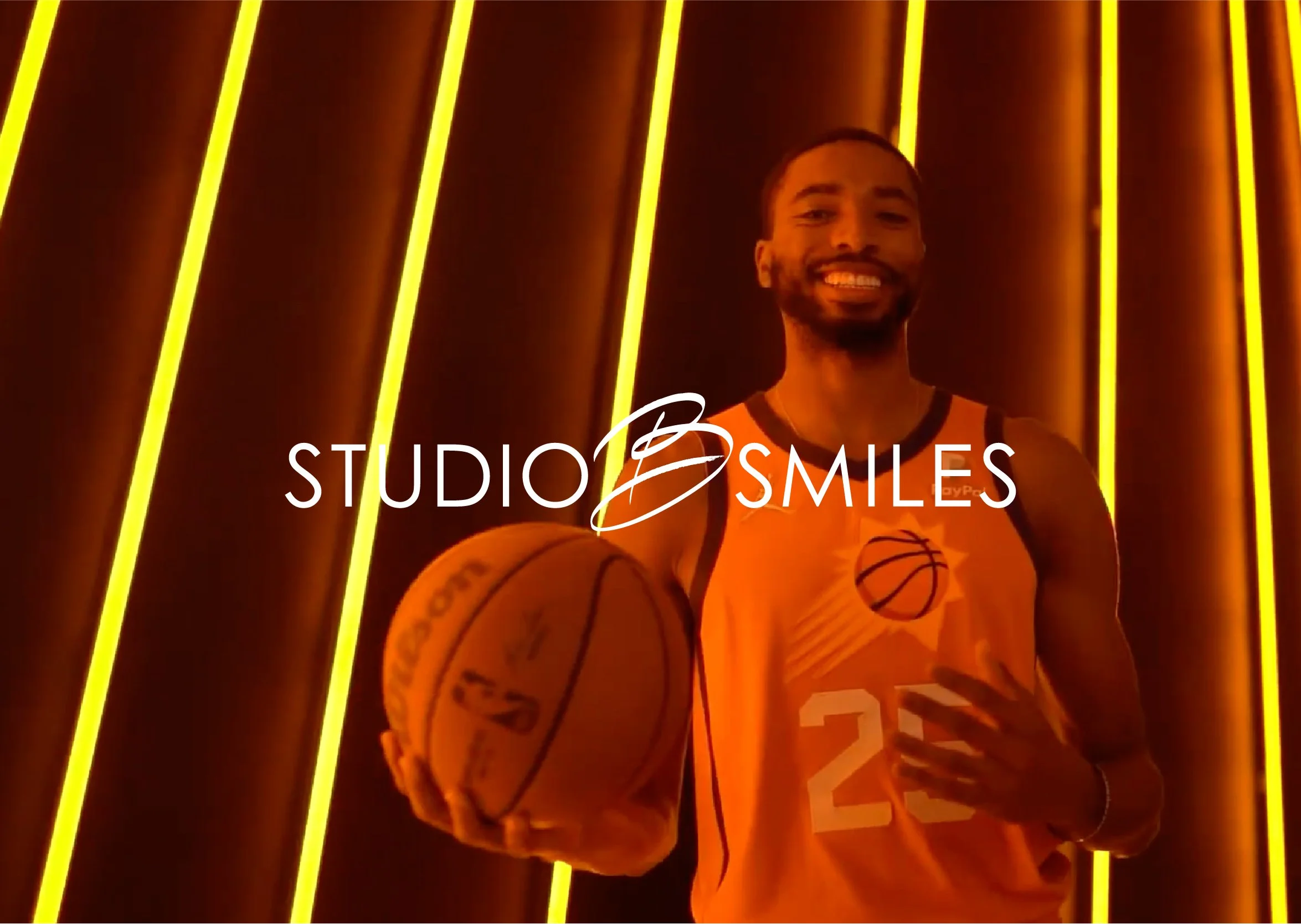 A smiling man wearing a basketball jersey holding a basketball, standing in front of vertical yellow neon lights with a dark background.