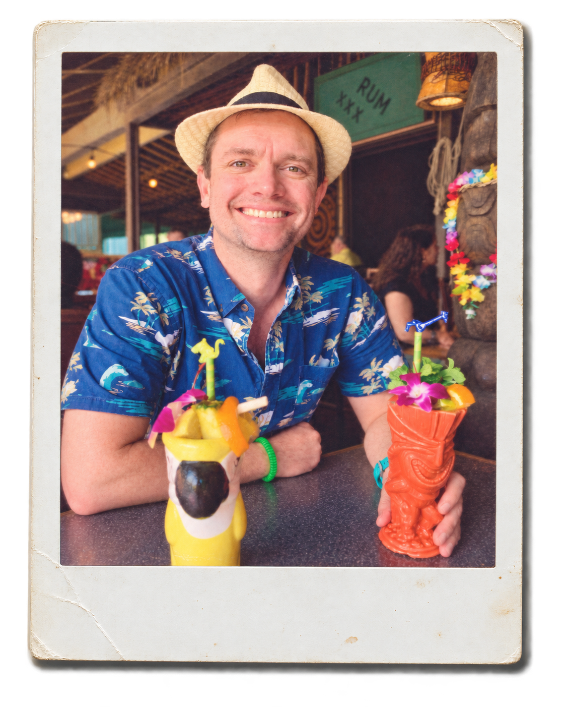 A man smiling at a tropical-themed bar, wearing a straw fedora, a blue Hawaiian shirt, and a green bracelet, sitting at a table with two colorful tiki-style drinks decorated with flowers and fruit.
