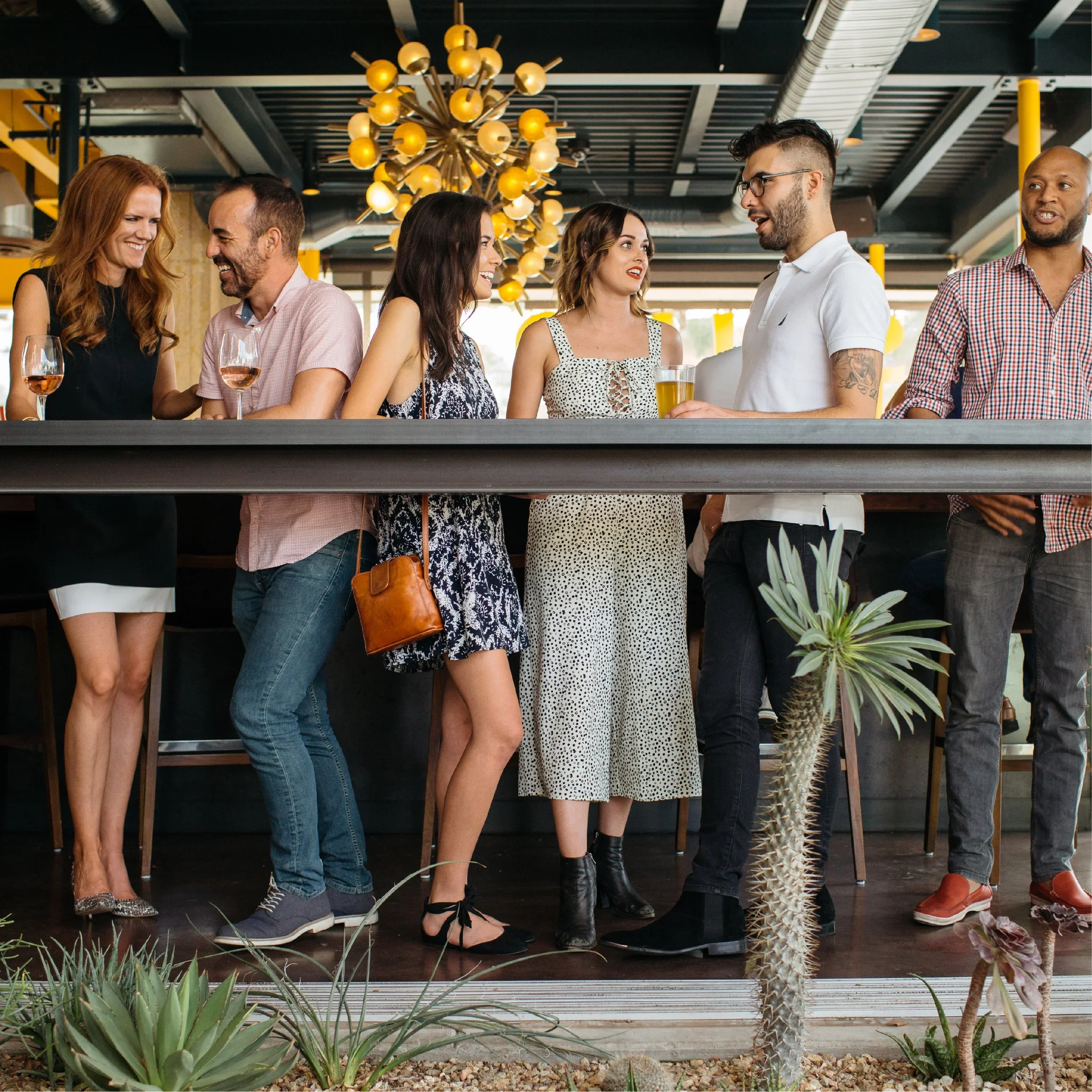A group of people socializing and enjoying drinks at a bar or restaurant with modern decor, including yellow and black elements and a large, decorative chandelier. There is greenery in the foreground.