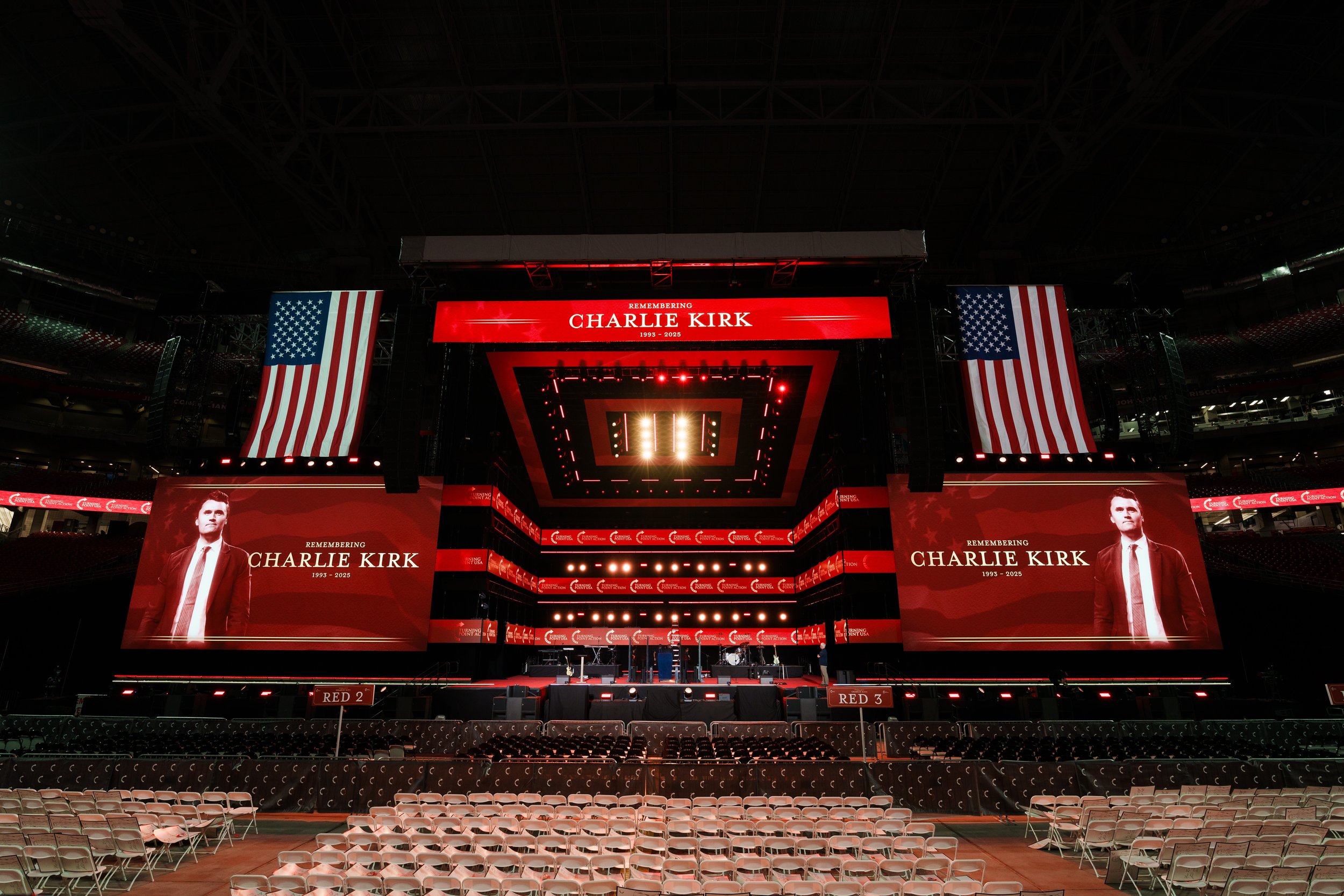Empty stadium with large screens displaying a tribute to Charlie Kirk, with American flags on either side and seating arranged below.