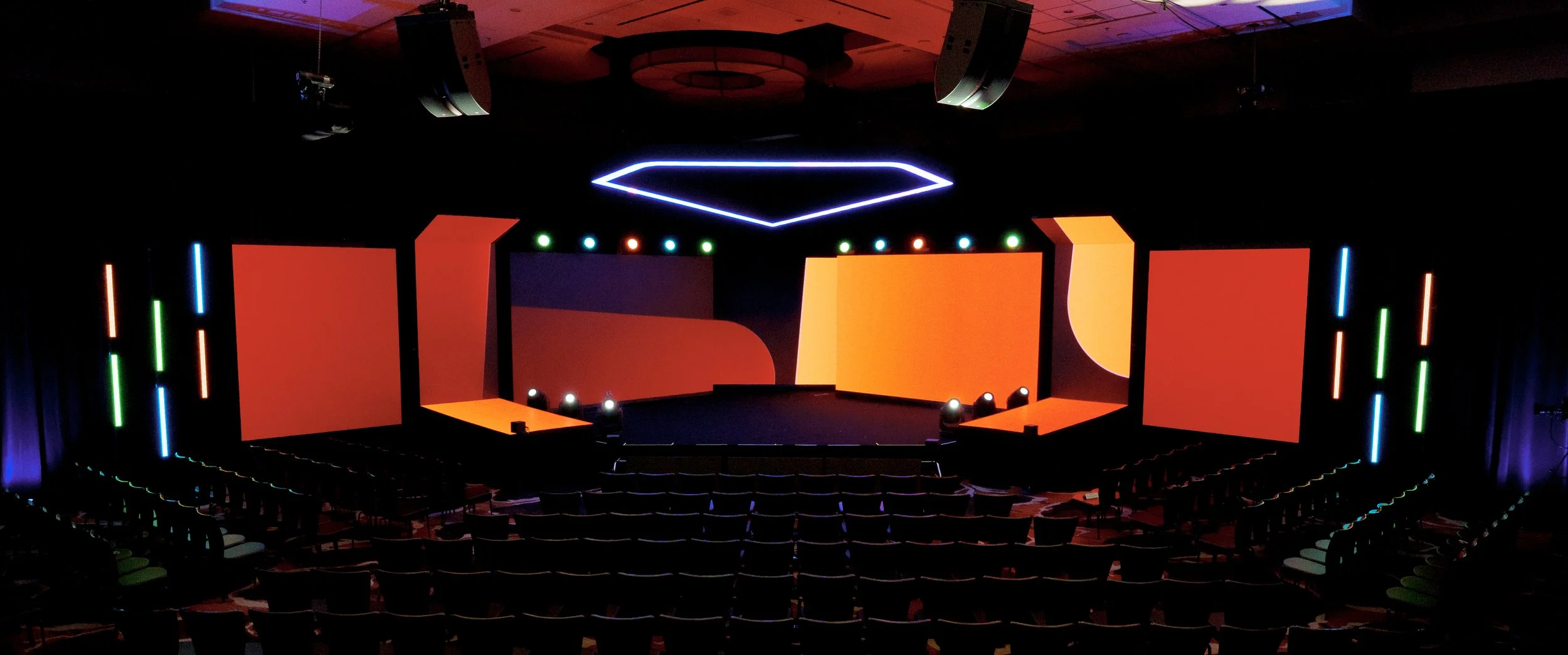 An empty auditorium stage with colorful LED lights, projection screens, and seating for the audience.