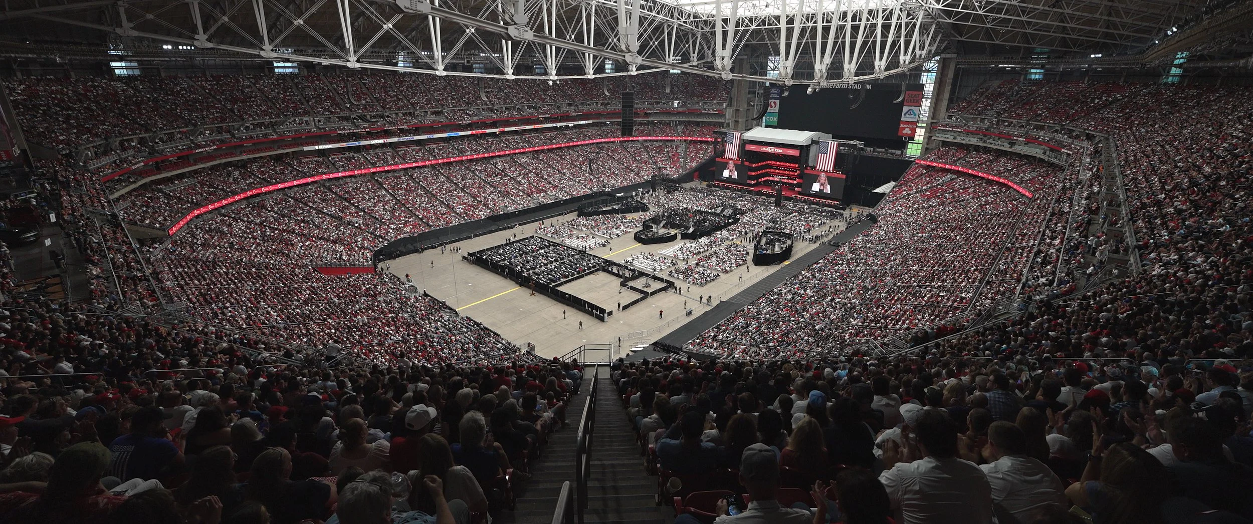 Inside a large indoor stadium filled with spectators seated in tiered bleachers for a concert or event, with a stage at the center and large screens displaying a speaker.