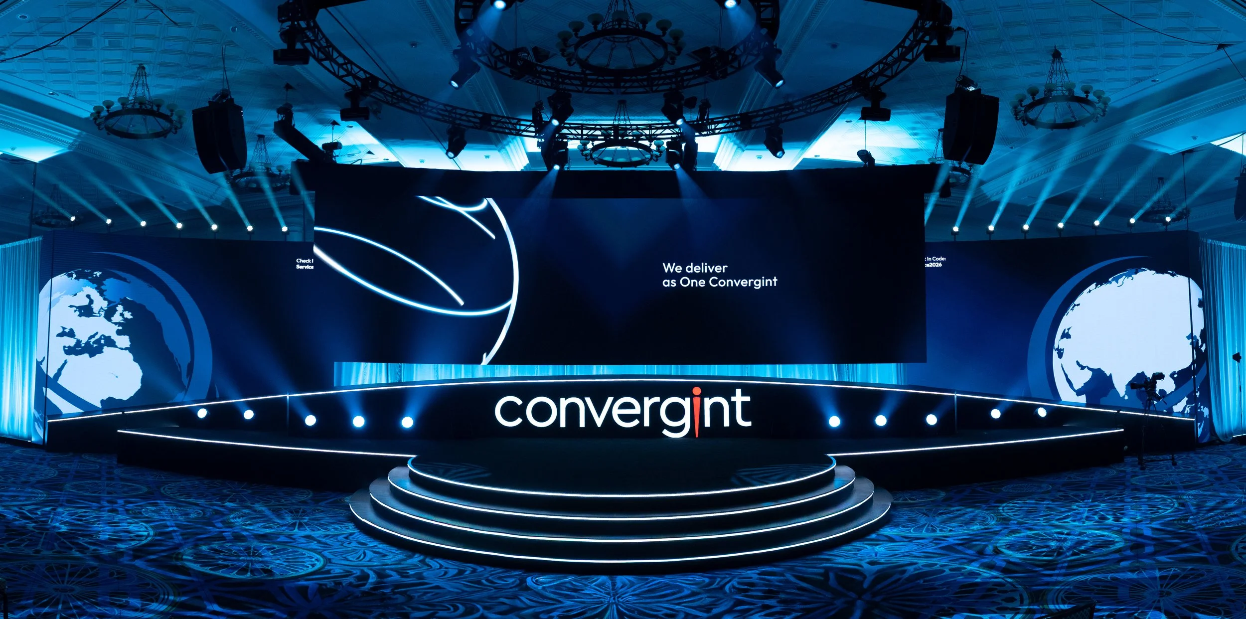 Conference stage with large screens displaying the text 'We deliver as One Convergint', a world map graphic, and the Convergint logo, with blue lighting and circular truss overhead.