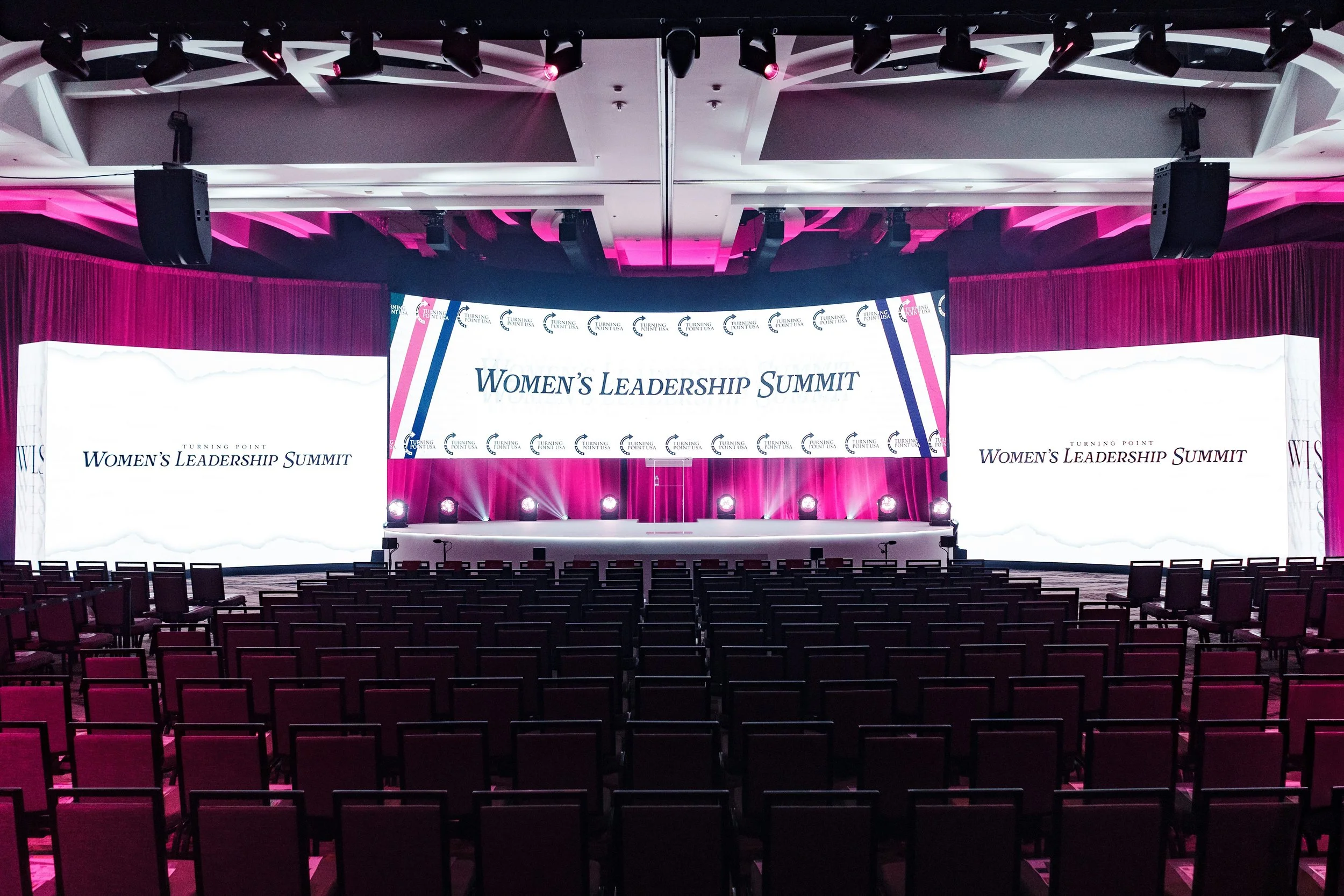 Stage set up for Women's Leadership Summit with large screens displaying event name, pink and purple lighting, and rows of chairs in front.
