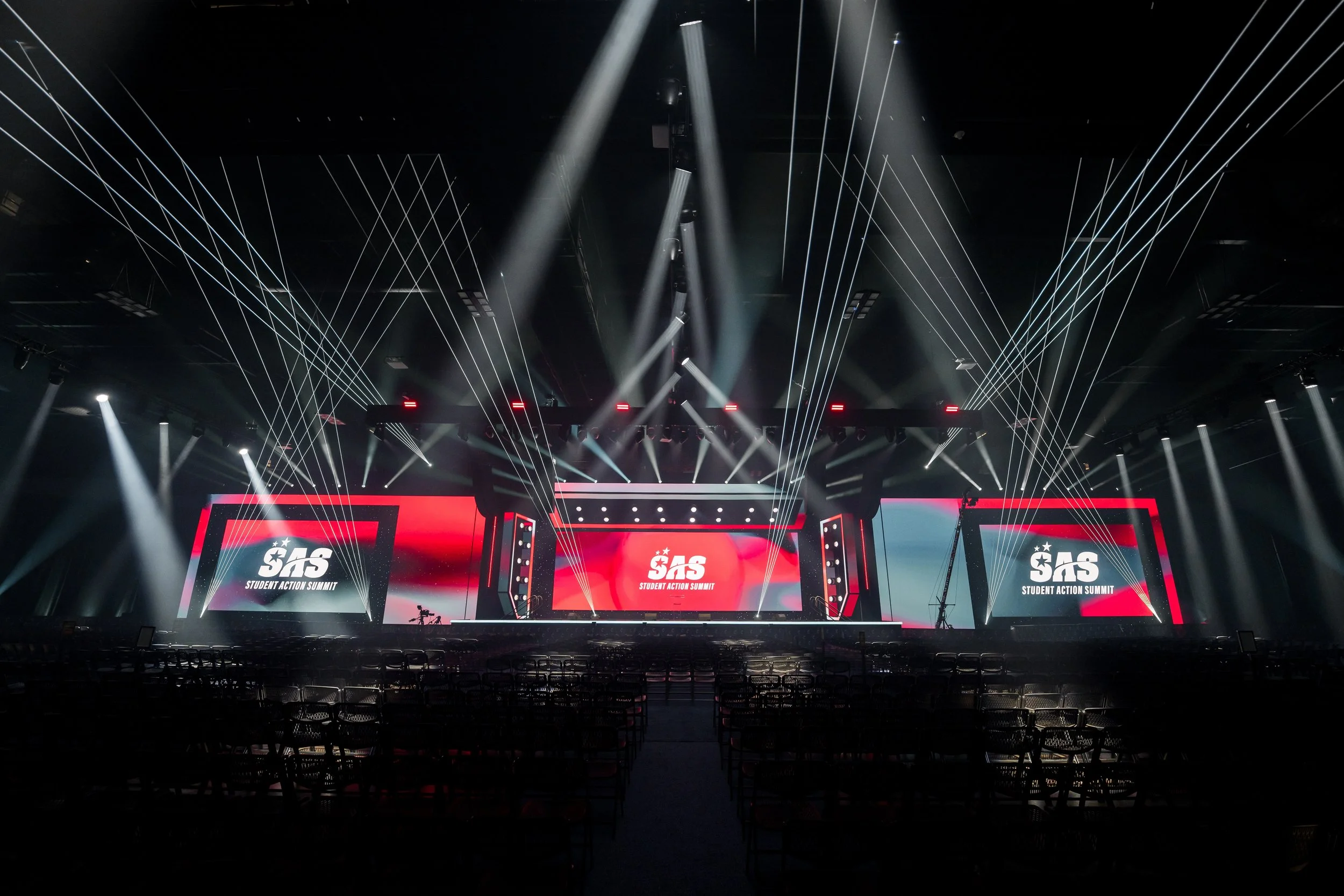 Concert stage with multiple large screens displaying the logo for SAS Student Action Summit, surrounded by multiple diagonal and vertical white stage lights and a dark auditorium with empty chairs.