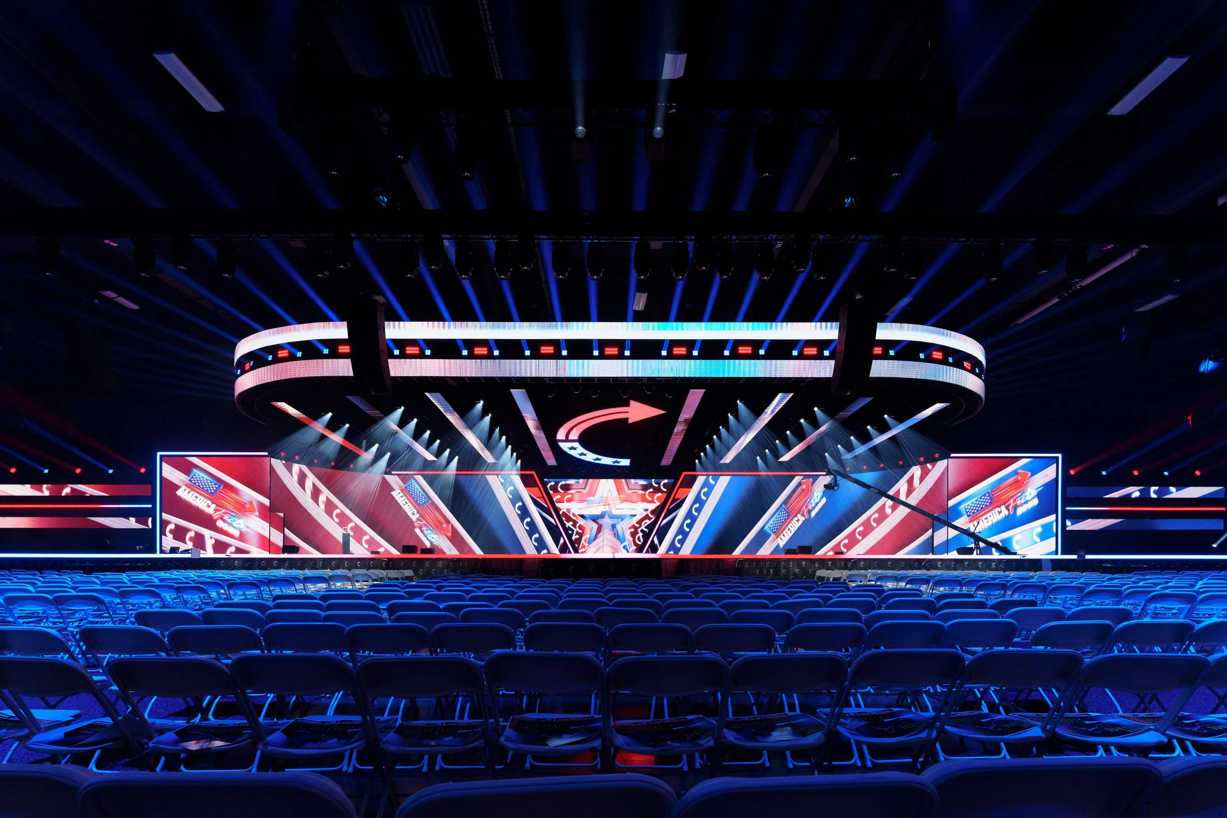 An empty stage with bright LED screens and overhead lighting, set up for a performance or event, with rows of blue chairs in front.