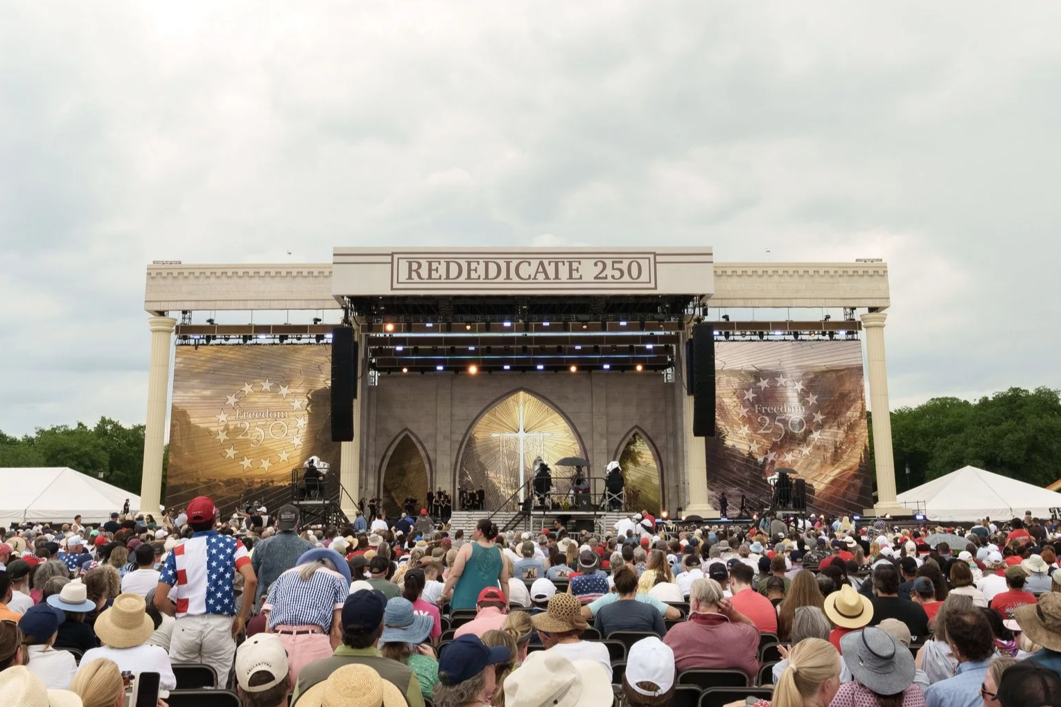 An outdoor stage with a large audience gathered to watch a concert, with banners celebrating the 250th anniversary of American independence and the word 'REDEDICATE 250' displayed above the stage.