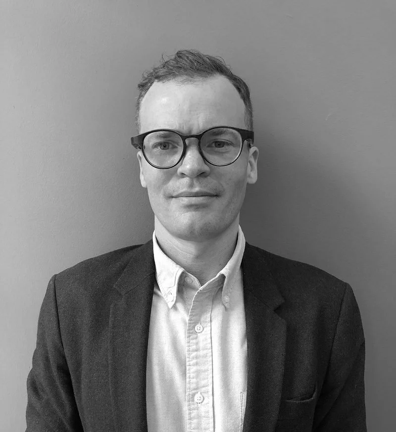 Black and white portrait of Marty McColl of Studio Zeal with short hair, wearing glasses, a white button-up shirt, and a dark blazer, standing against a plain wall.