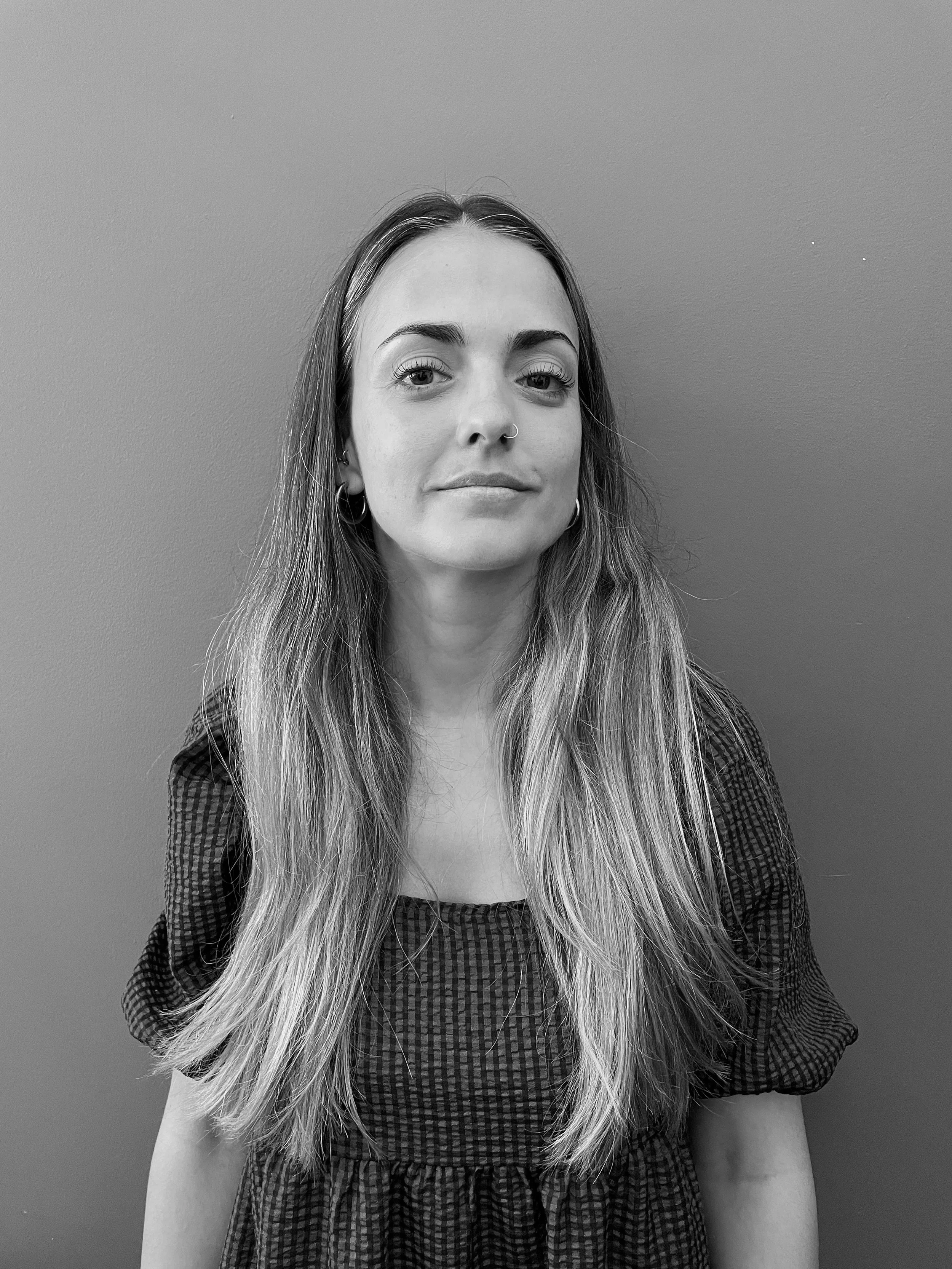 Black and white portrait of Diana Cirne of Studio Zeal with long wavy hair, wearing a checkered dress, standing against a plain wall.