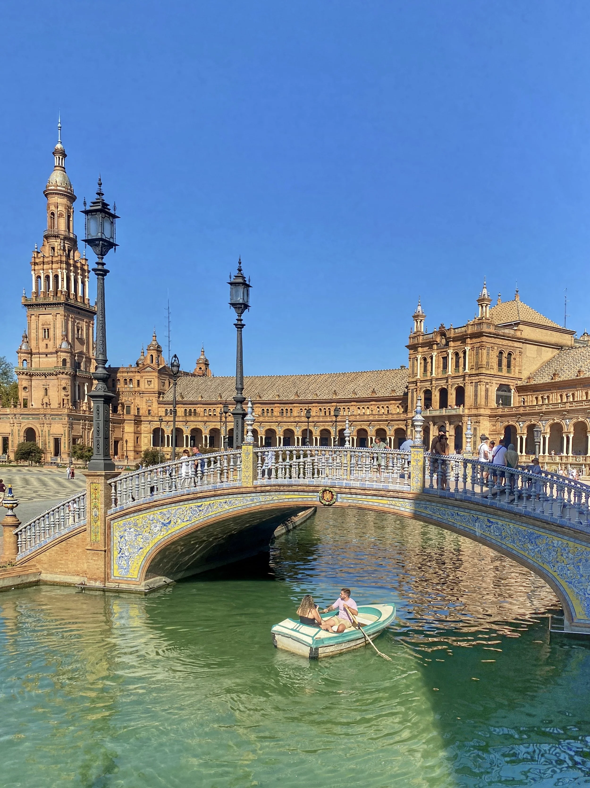A man rows a boat with a woman in it in the moat of the Plaza de España, heading under a curved bridge
