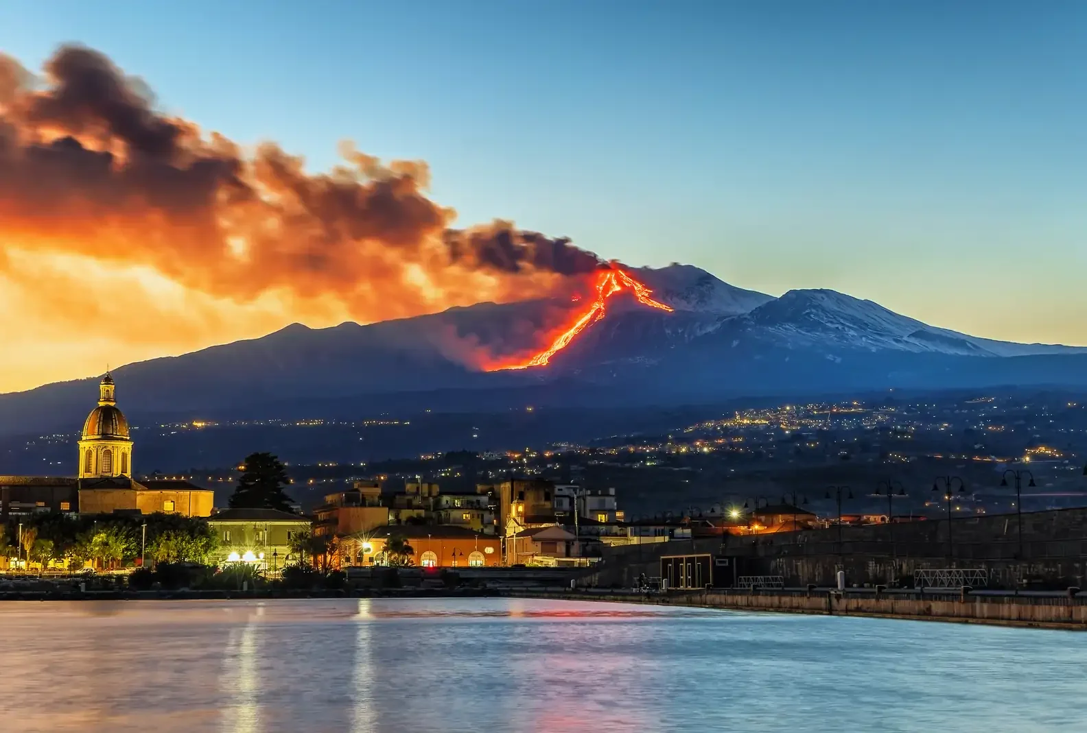Mount Etna erupting, with smoke and lava on Sicily, Italy, with a city in the foreground in the evening